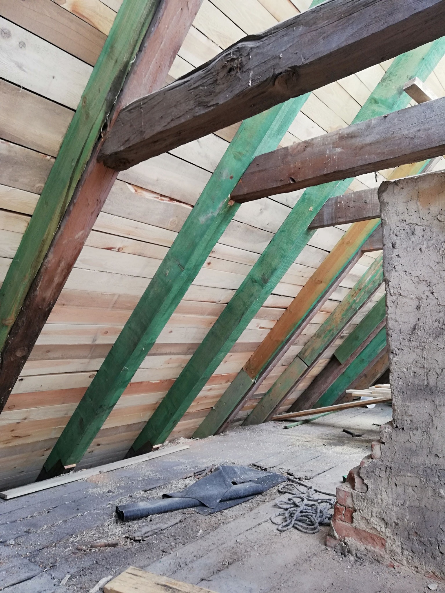 Surowe drewno więźby dachowej z zielonymi elementami konstrukcyjnymi, widoczne deski poszycia i fragment starego muru. Ujęcie z dołu, w trakcie prac.
