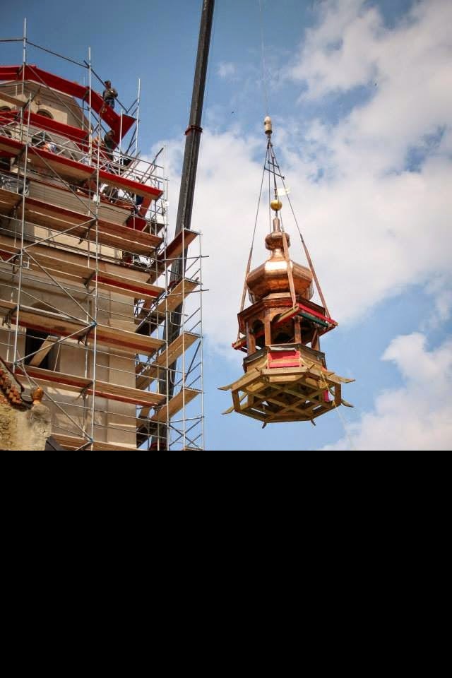 Montaż kopuły wieży kościelnej za pomocą dźwigu. Na pierwszym planie kopuła z miedzianym dachem i drewnianą konstrukcją. W tle rusztowania i fragment budynku.