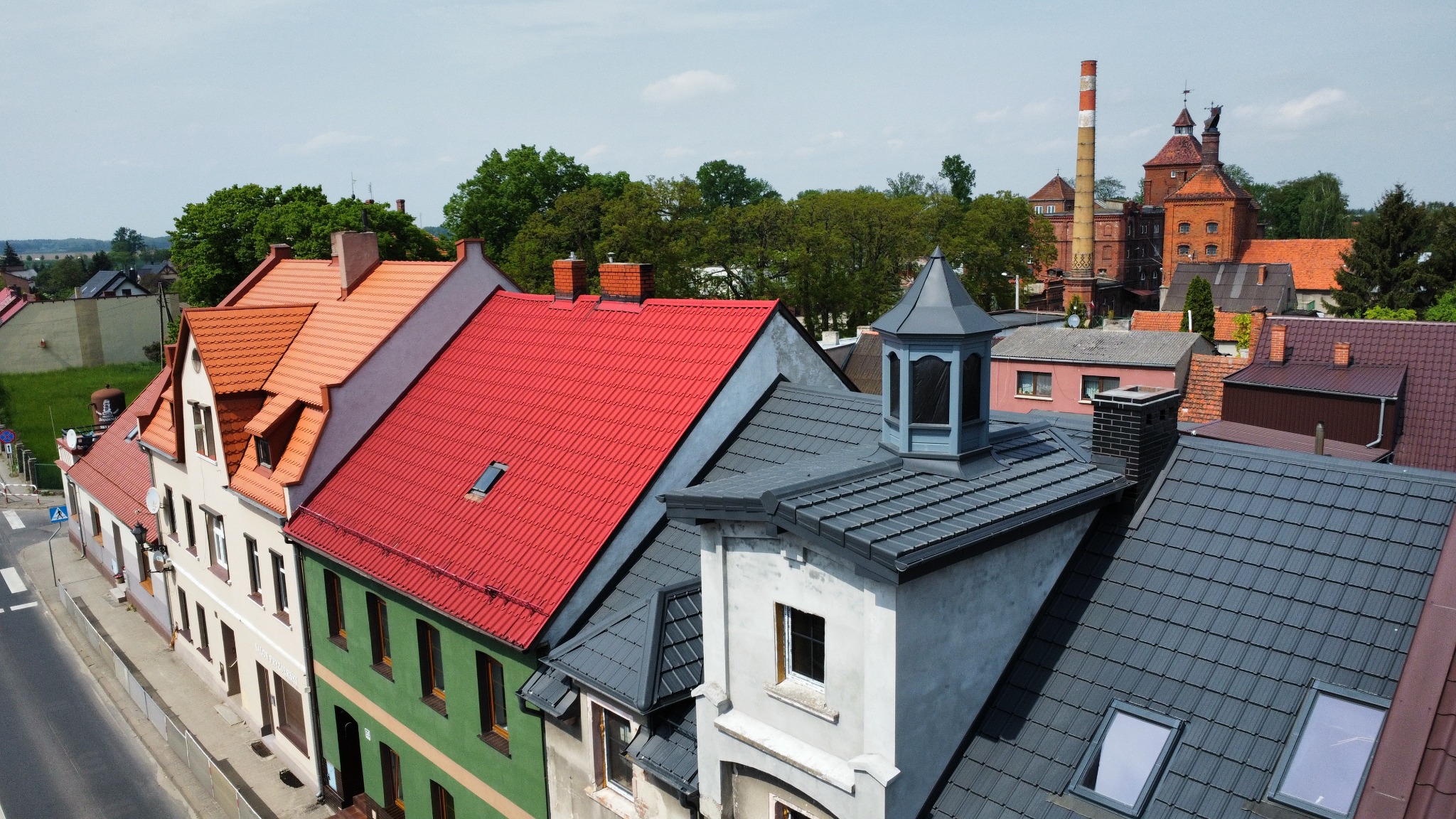 Widok z góry na szereg budynków z dachami w odcieniach czerwieni i szarości, z kominami i lukarnami. W tle ceglany budynek z kominem. Słoneczny dzień.