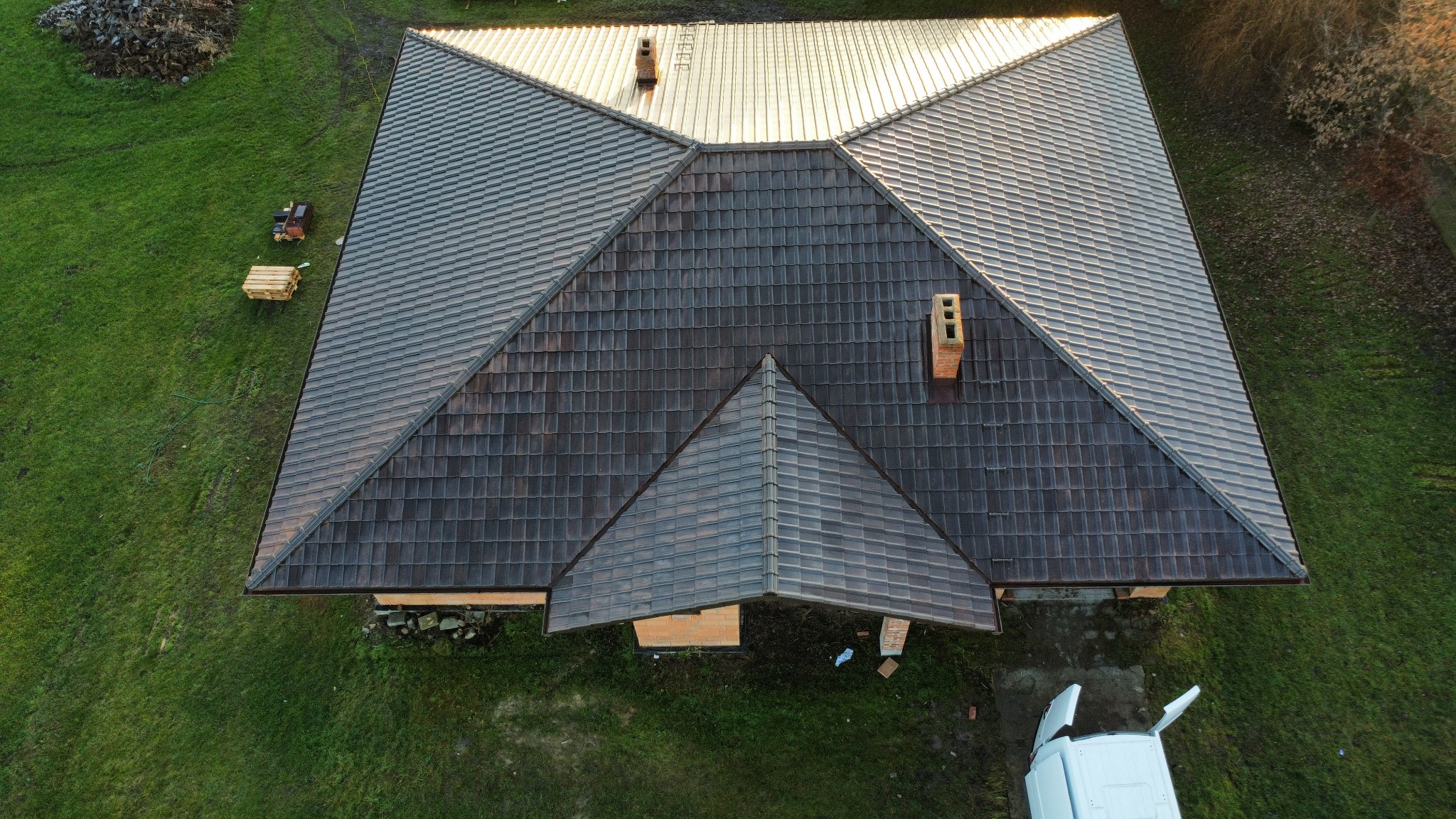 Widok z góry na brązowy dach z dachówki ceramicznej, z kominem i elementami obróbki blacharskiej. Obok dachu leżą palety z materiałami budowlanymi.