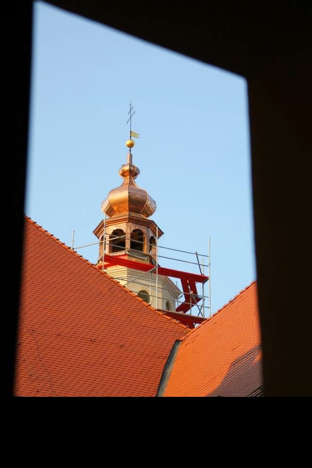 Miedziana kopuła wieży z rusztowaniem, widziana zza okna. Czerwona dachówka w dolnej części kadru, błękitne niebo w tle. Detal architektoniczny w trakcie renowacji.