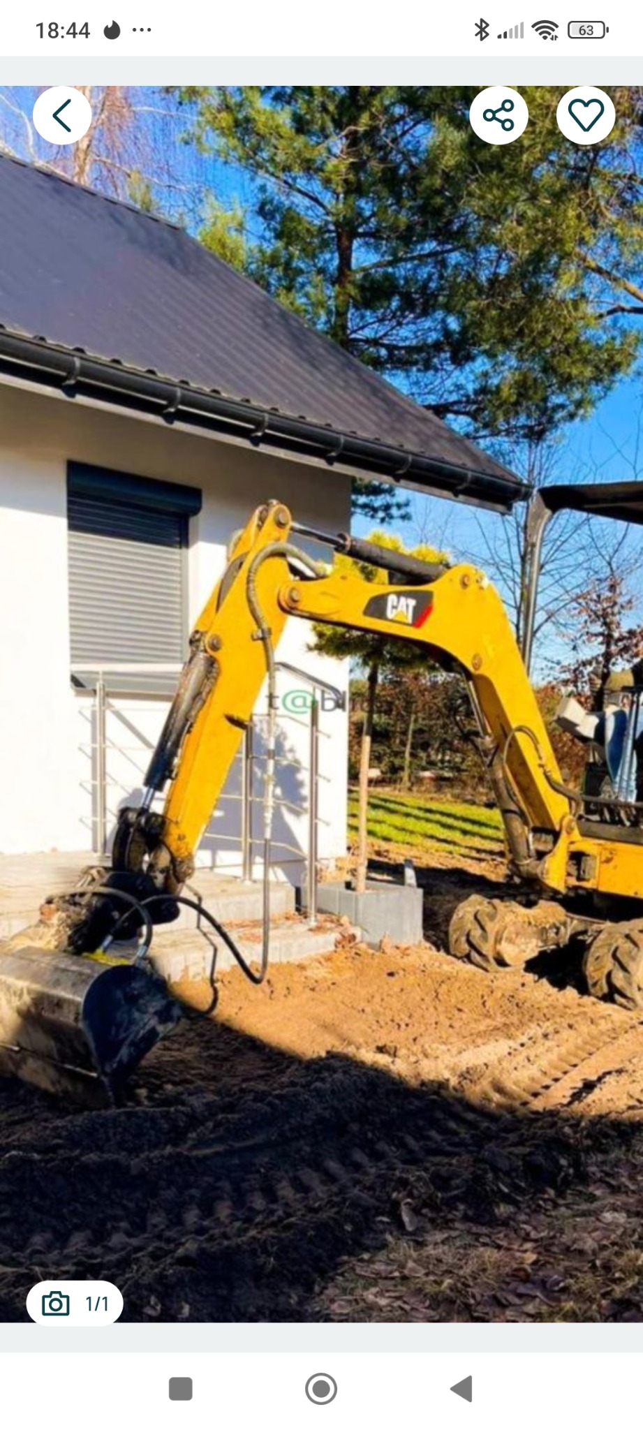 Żółta koparka CAT wykonuje wykop obok budynku z szarymi roletami i metalową balustradą. Ślady opon na ziemi. Błękitne niebo w tle.