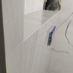 Oleksandr - Ściana wyłożona jasnymi, marmurkowymi płytkami. Widoczny fragment instalacji i narzędzie glazurnicze. Ujęcie z bliska, ukazujące precyzję wykonania.