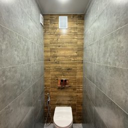 Oleksandr - Wąska łazienka po remoncie w Kaliszu. Szare płytki imitujące beton na ścianach kontrastują z drewnopodobnymi płytkami za toaletą. Widoczny bidet i minimalistyczny design.
