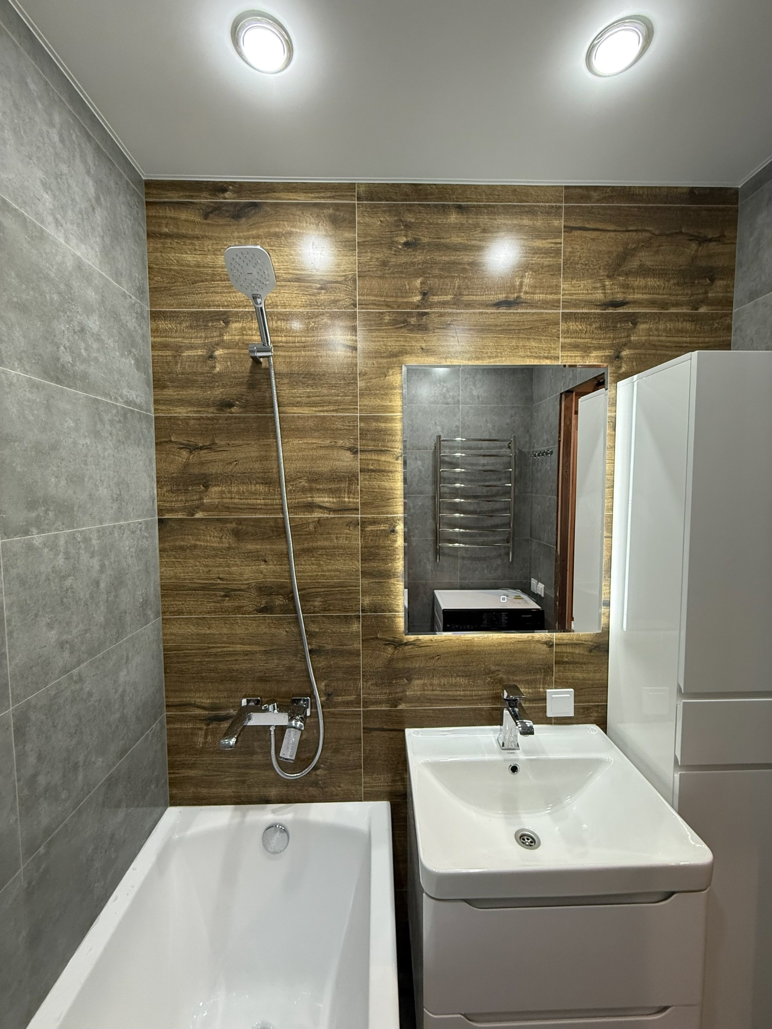 Nowoczesna łazienka z drewnopodobnymi płytkami, białą armaturą i podświetlanym lustrem. Widok na wannę, umywalkę z szafką i grzejnik w odbiciu lustra.