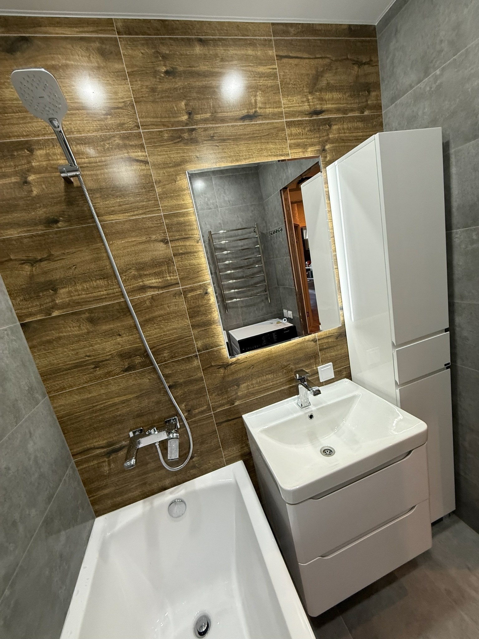 Nowoczesna łazienka po remoncie: drewnopodobne płytki, biała armatura (wanna, umywalka z szafką, słupek), lustro z odbiciem grzejnika drabinkowego.