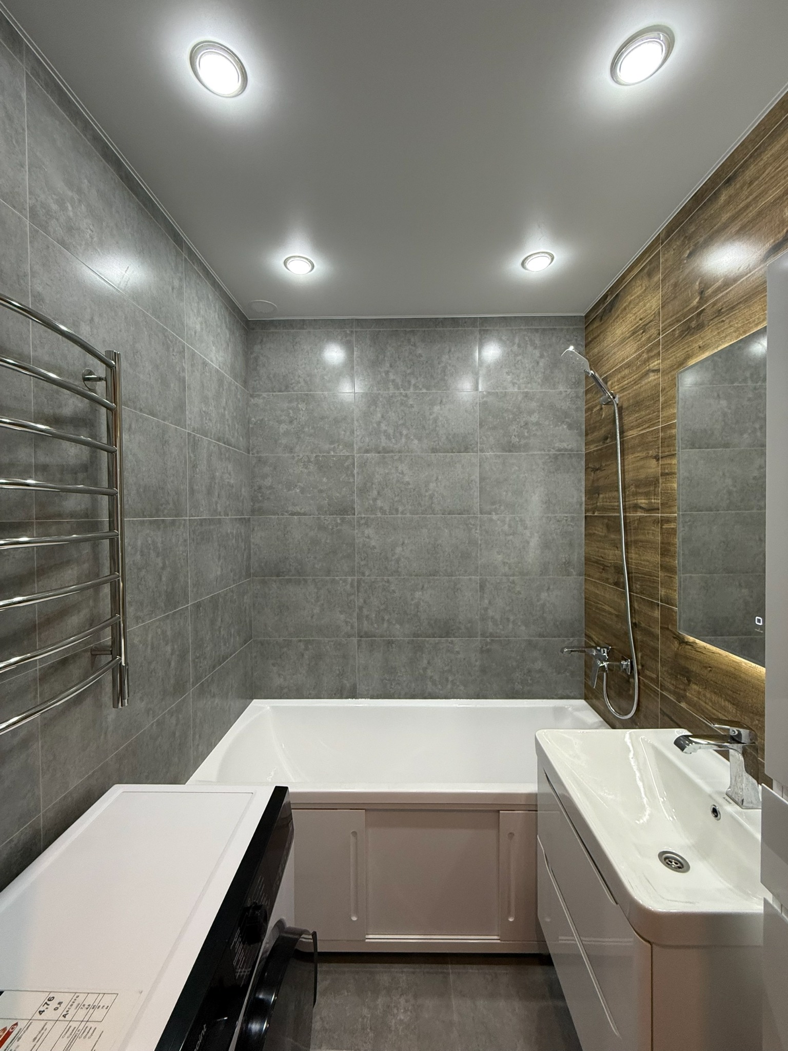 Nowoczesna łazienka z szarymi płytkami, drewnopodobną ścianą, wanną z obudową, umywalką z szafką i pralką. Oświetlenie sufitowe punktowe.