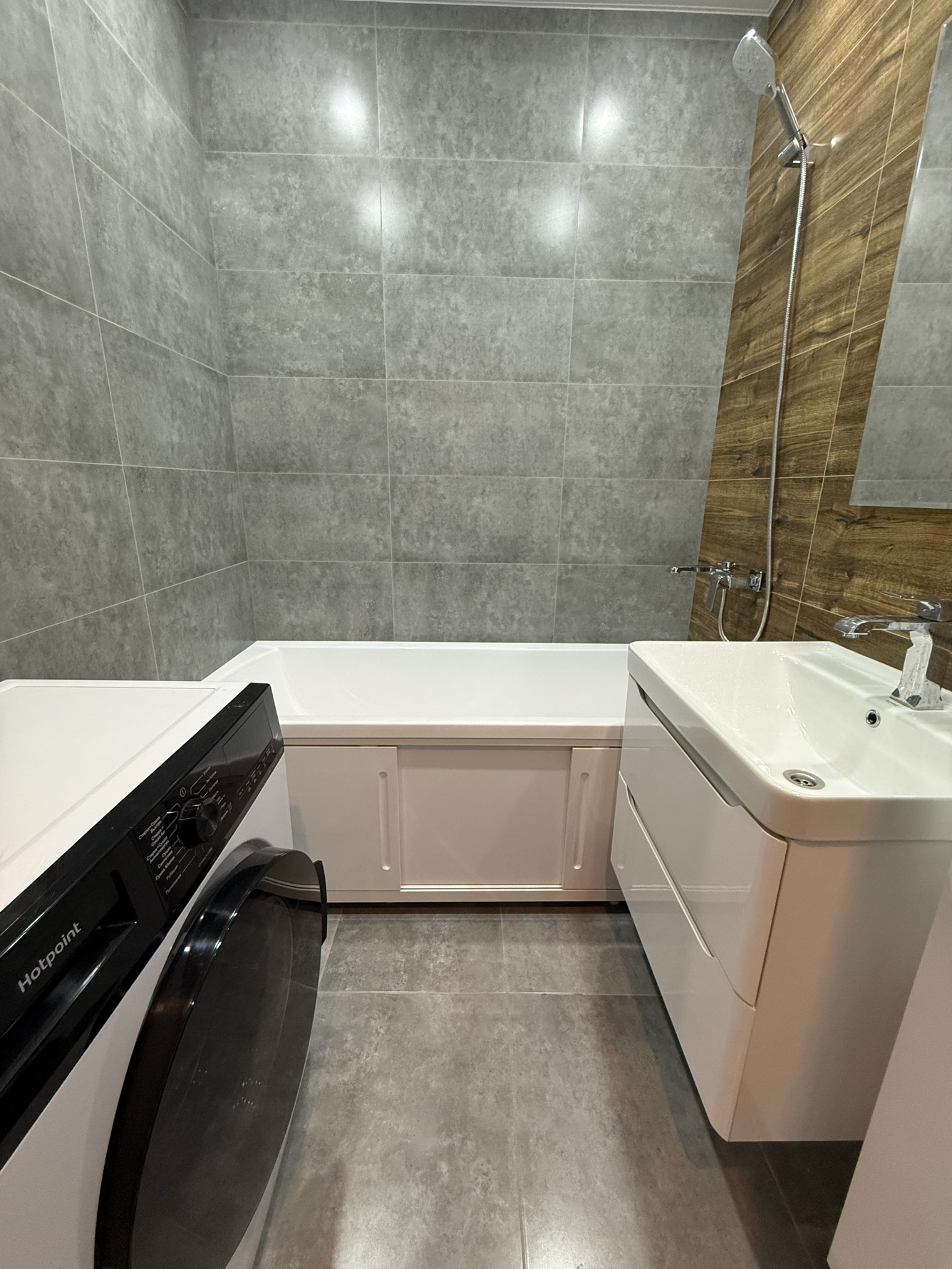 Nowoczesna łazienka z szarymi płytkami, białą wanną obudowaną panelami, umywalką z szafką i pralką Hotpoint. Minimalistyczny design, drewnopodobne akcenty.