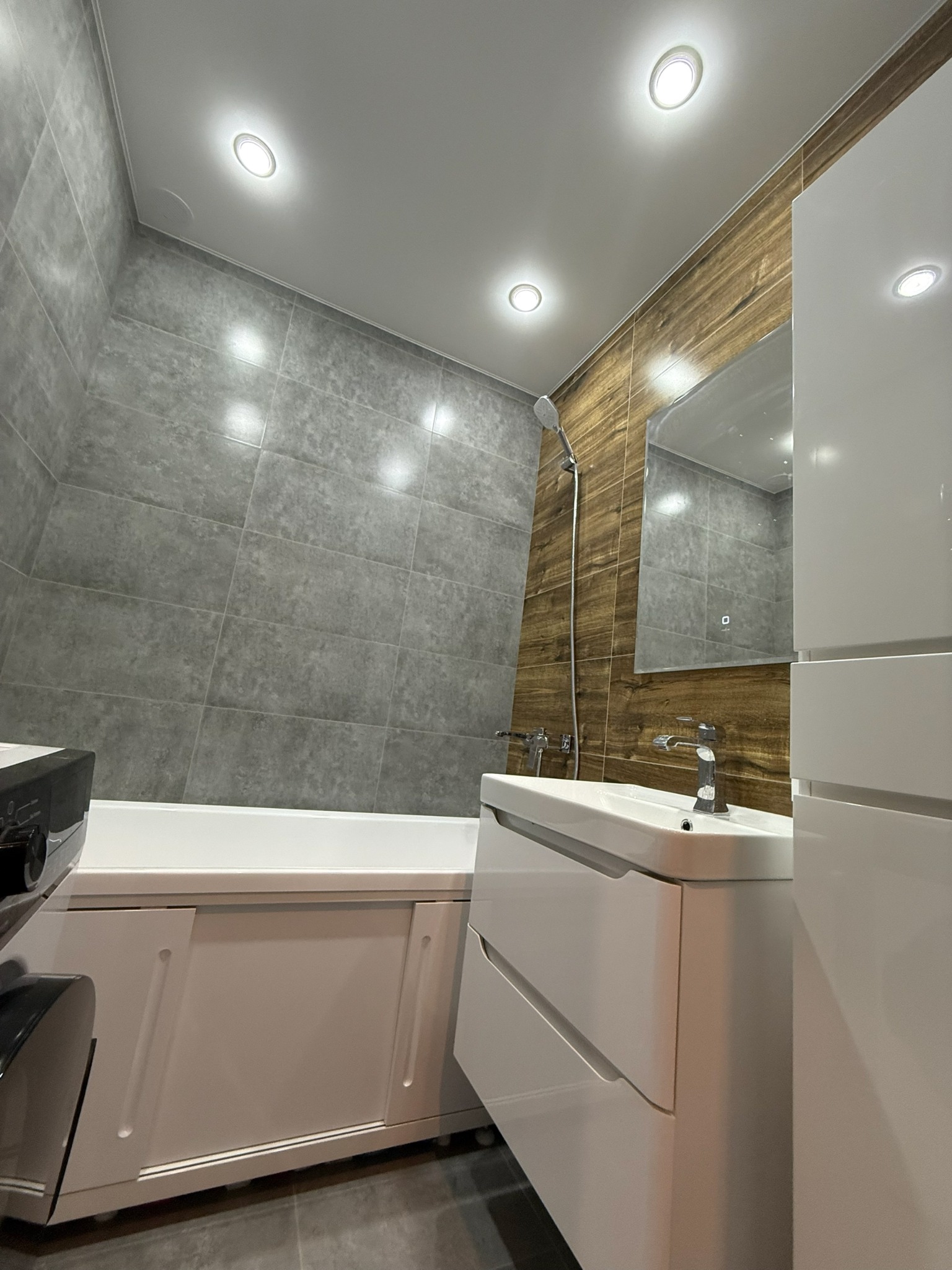 Nowoczesna łazienka po remoncie: szare płytki, drewnopodobne akcenty, biała armatura i oświetlenie punktowe. Minimalistyczny design z dbałością o detale.