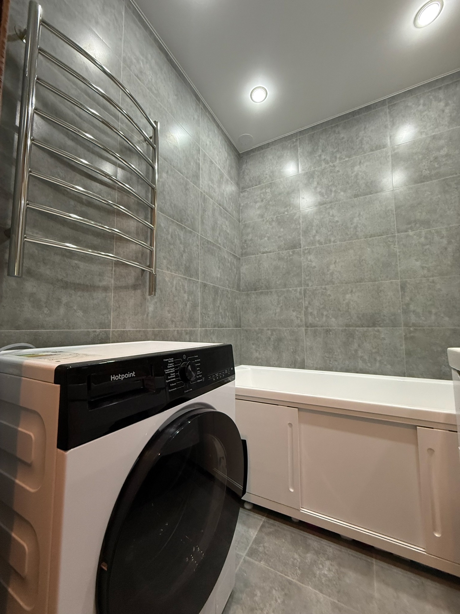 Szara łazienka z pralką Hotpoint, grzejnikiem drabinkowym i zabudowaną wanną z przesuwnymi drzwiami. Minimalistyczny design, płytki imitujące kamień.