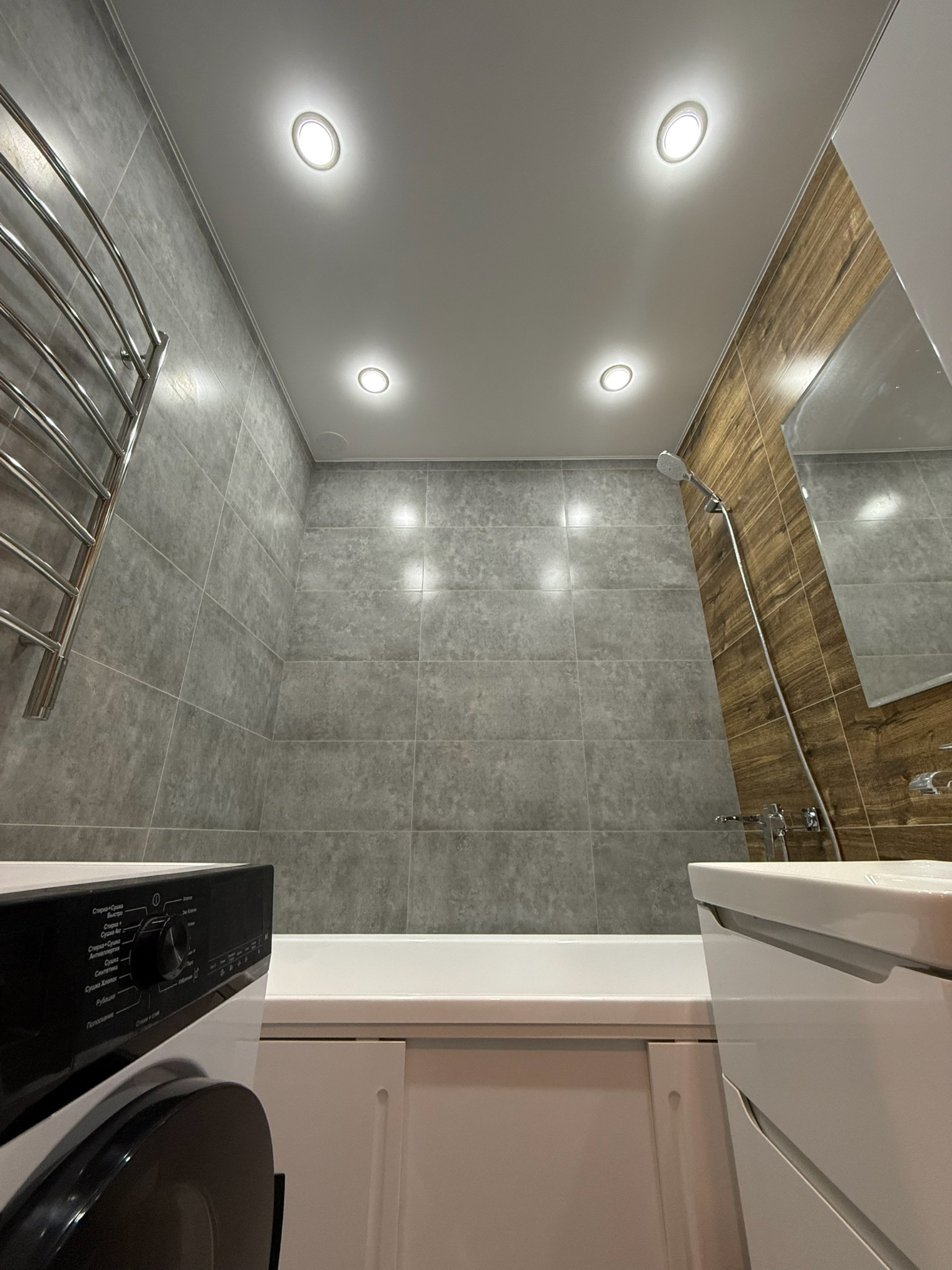 Nowoczesna łazienka z szarymi płytkami, drewnopodobnymi akcentami i podwieszanym sufitem z oświetleniem punktowym. Widoczna wanna, umywalka i pralka.