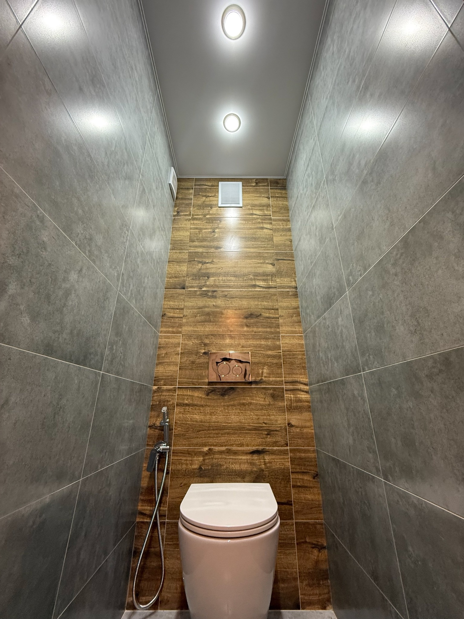Wąska toaleta z szarymi płytkami i drewnopodobną ścianą akcentującą. Bidetka obok sedesu. Minimalistyczne oświetlenie sufitowe.