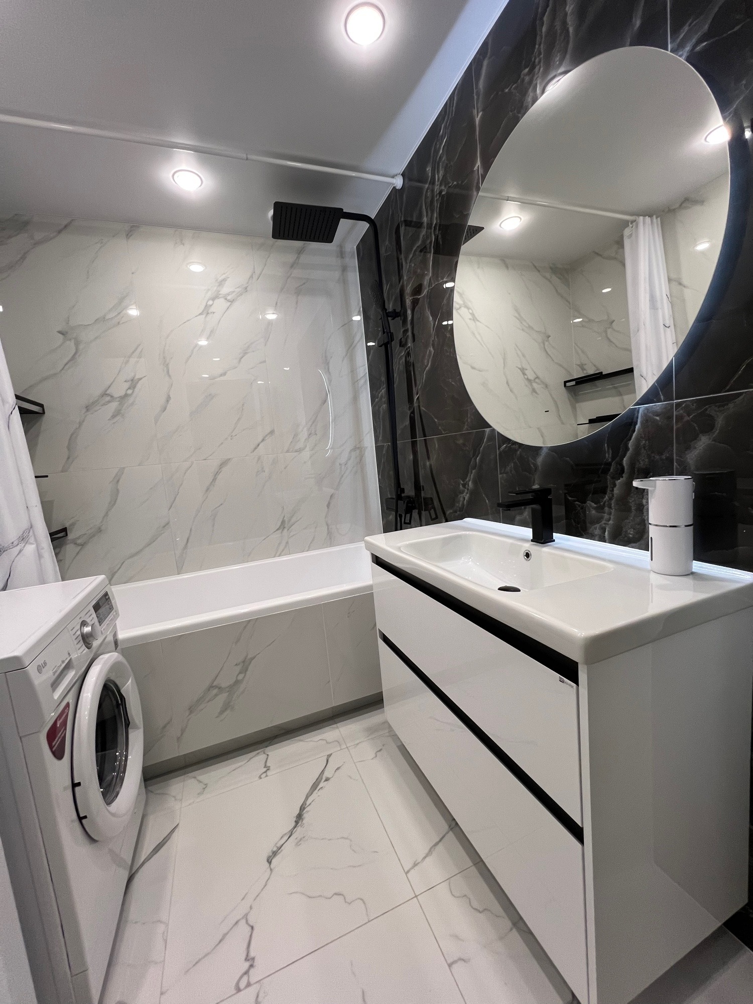 Nowoczesna łazienka z czarno-białymi marmurowymi płytkami, owalnym lustrem z podświetleniem, białą umywalką z szafką i pralką LG. Minimalistyczny design.