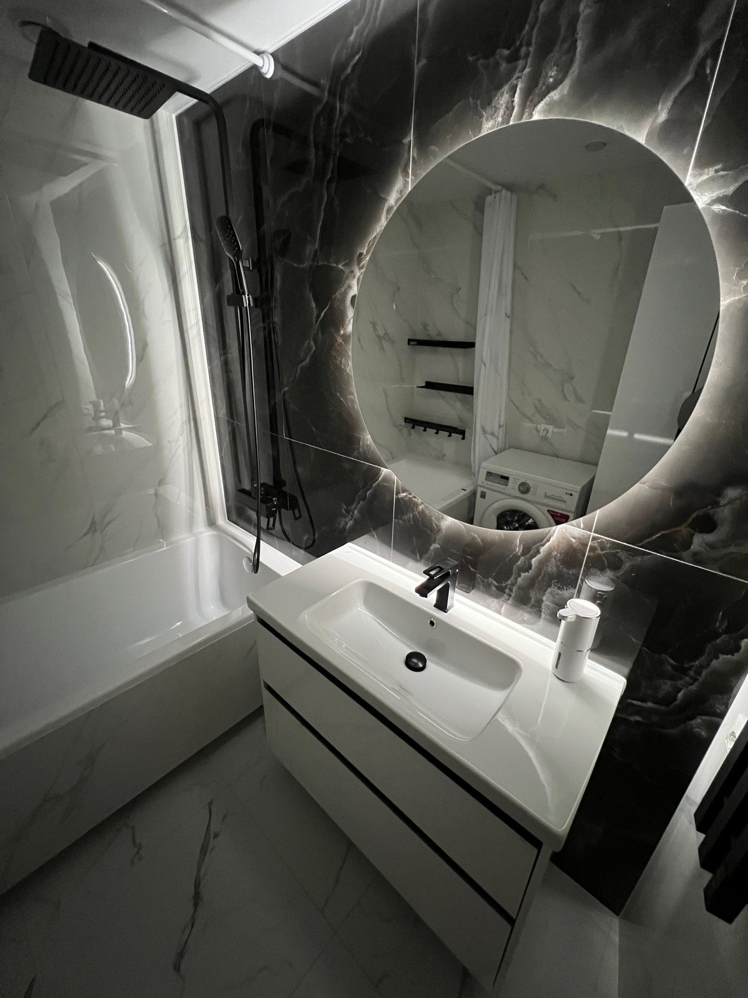 Nowoczesna łazienka z czarno-białymi marmurowymi płytkami, okrągłym lustrem z podświetleniem LED i czarną armaturą. W odbiciu lustra widoczna pralka.