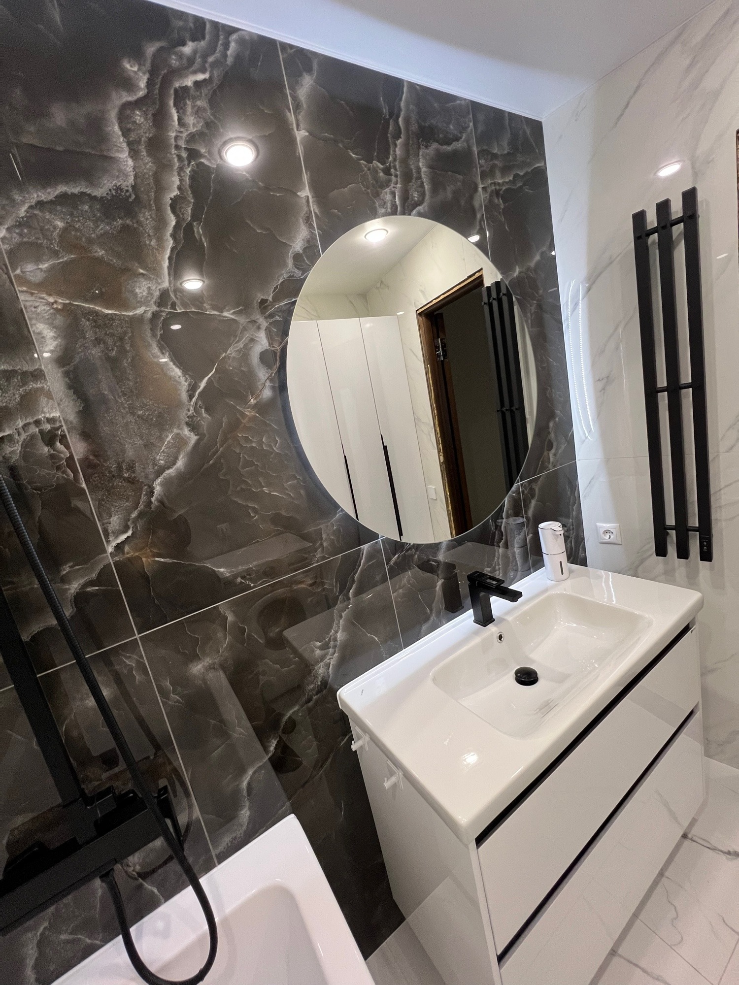 Nowoczesna łazienka z czarno-białymi płytkami imitującymi marmur, okrągłym lustrem i białą szafką pod umywalką z czarną armaturą. Widoczny grzejnik drabinkowy.