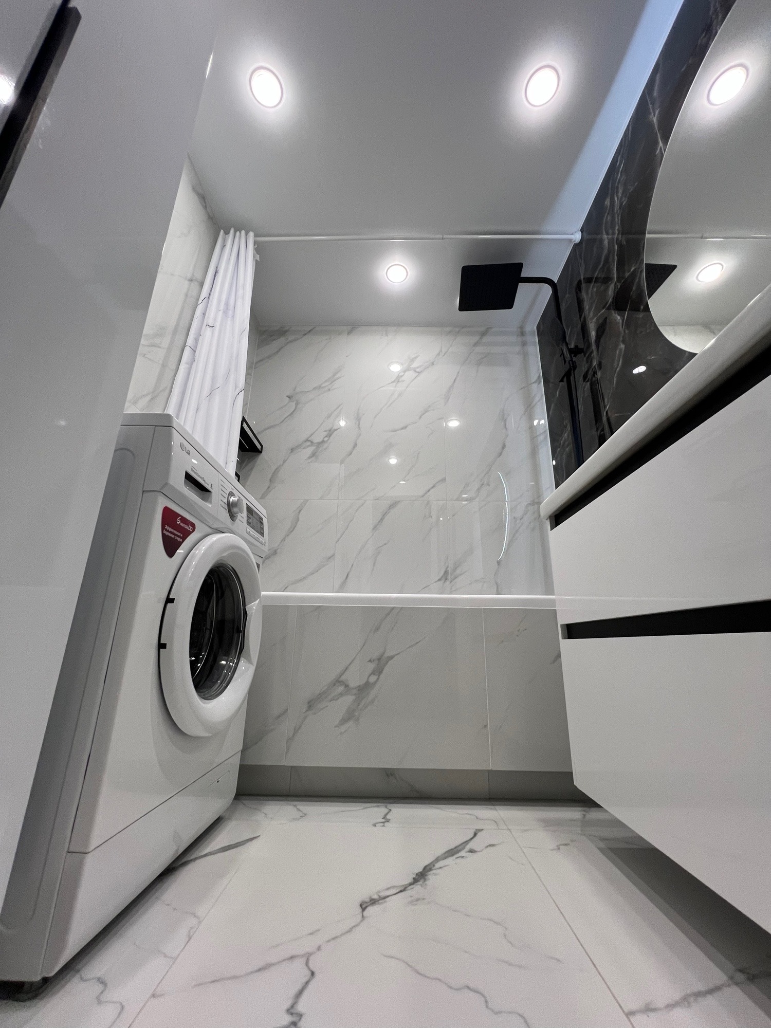 Nowoczesna łazienka z białą pralką LG, marmurowymi płytkami i czarnymi akcentami armatury. Minimalistyczny design, oświetlenie punktowe.
