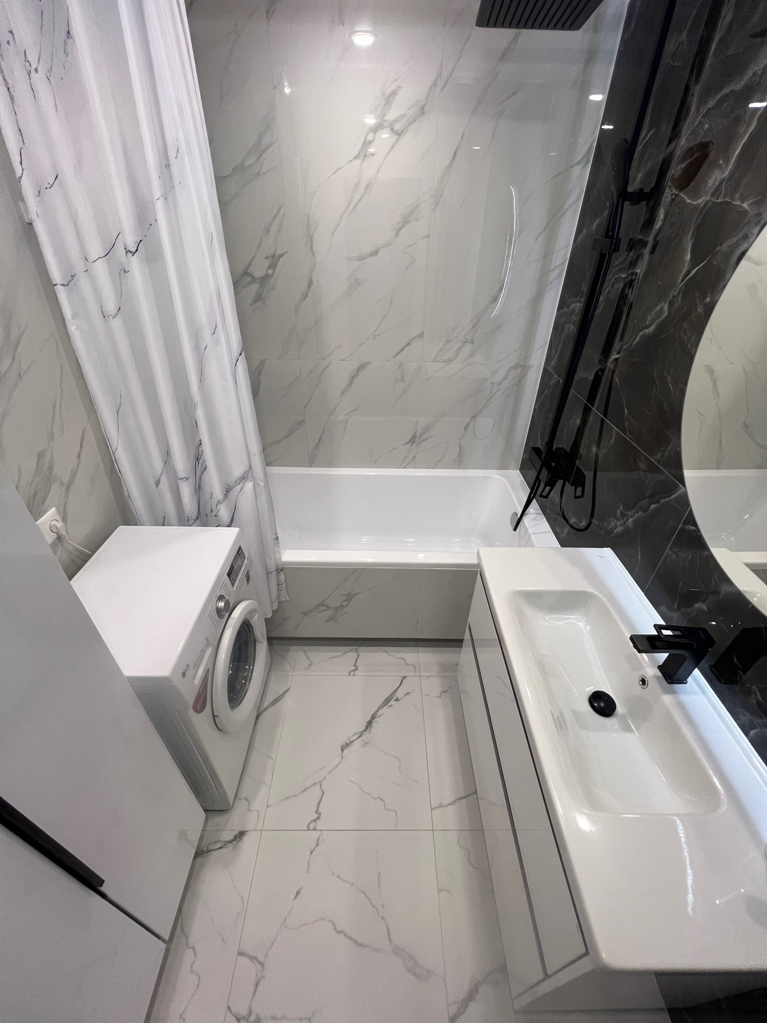 Nowoczesna łazienka z marmurowymi płytkami, białą armaturą i czarnymi akcentami. Widoczna pralka, wanna i umywalka z czarną baterią. Minimalistyczny design.