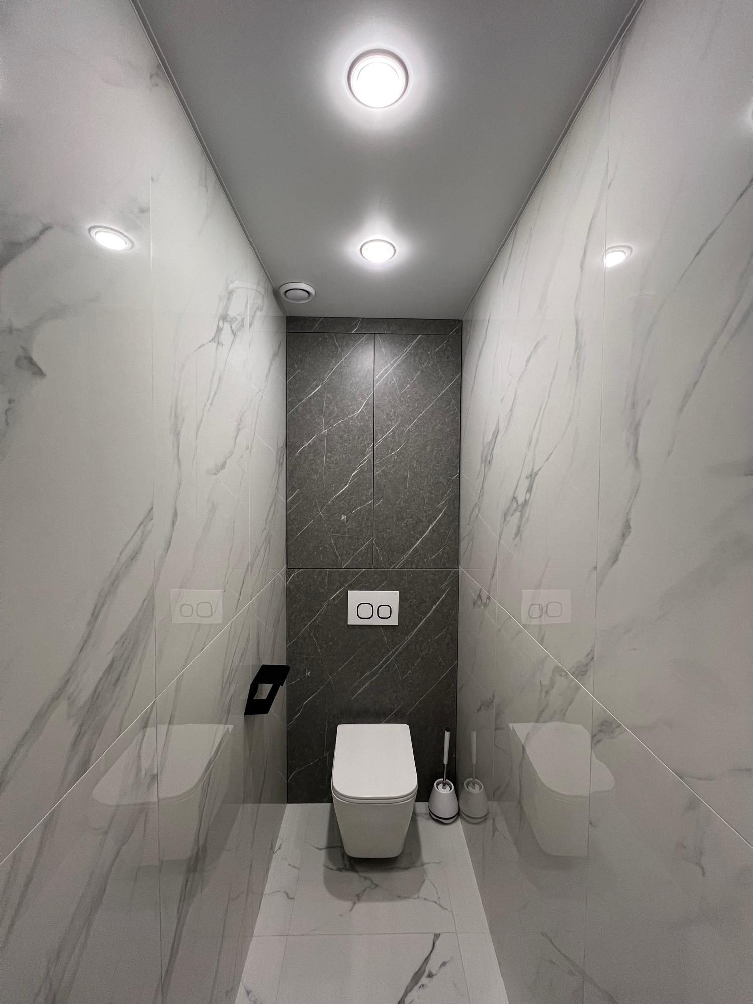Wąska łazienka z marmurowymi płytkami, podwieszaną toaletą i zabudową w kolorze grafitu. Minimalistyczny design z oświetleniem punktowym.