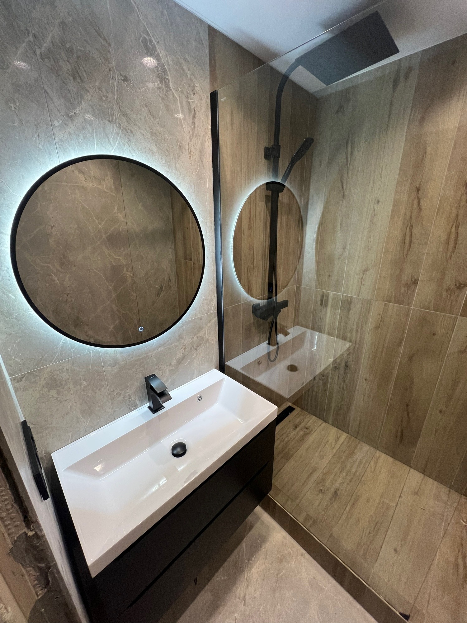 Nowoczesna łazienka z okrągłym lustrem LED, czarną armaturą i kabiną prysznicową typu walk-in. Płytki imitujące drewno i marmur. Minimalistyczny design.