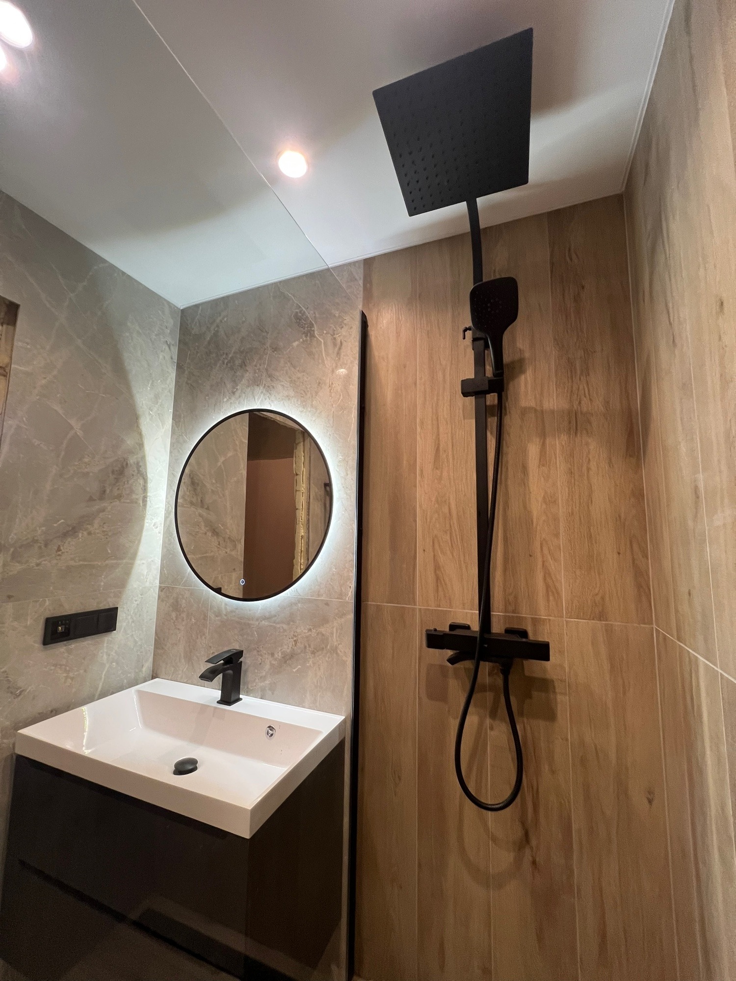 Nowoczesna łazienka z okrągłym lustrem LED, czarną armaturą i płytkami imitującymi drewno i marmur. Minimalistyczny design, oświetlenie punktowe.