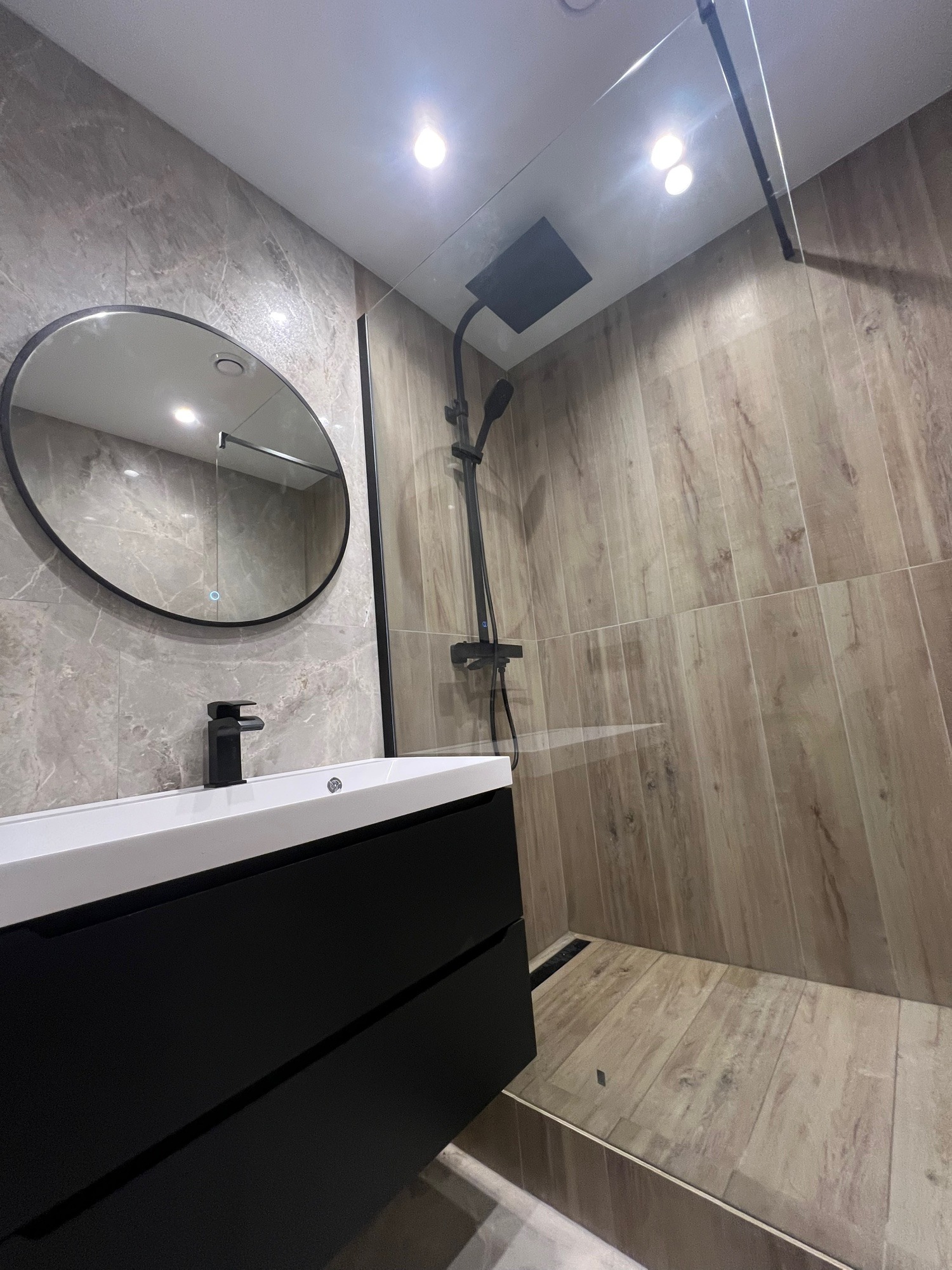 Nowoczesna łazienka z kabiną typu walk-in, umywalką nablatową na czarnej szafce i okrągłym lustrem z podświetleniem. Płytki imitujące drewno i marmur.