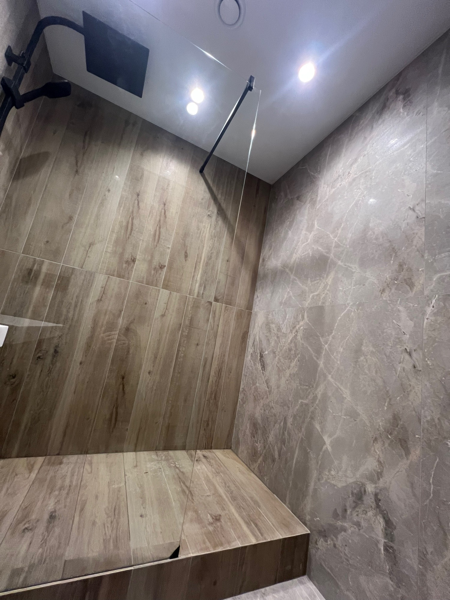 Nowoczesna kabina prysznicowa z drewnopodobnymi płytkami i szarą marmurową ścianą. Czarna armatura i oświetlenie punktowe podkreślają minimalistyczny design.