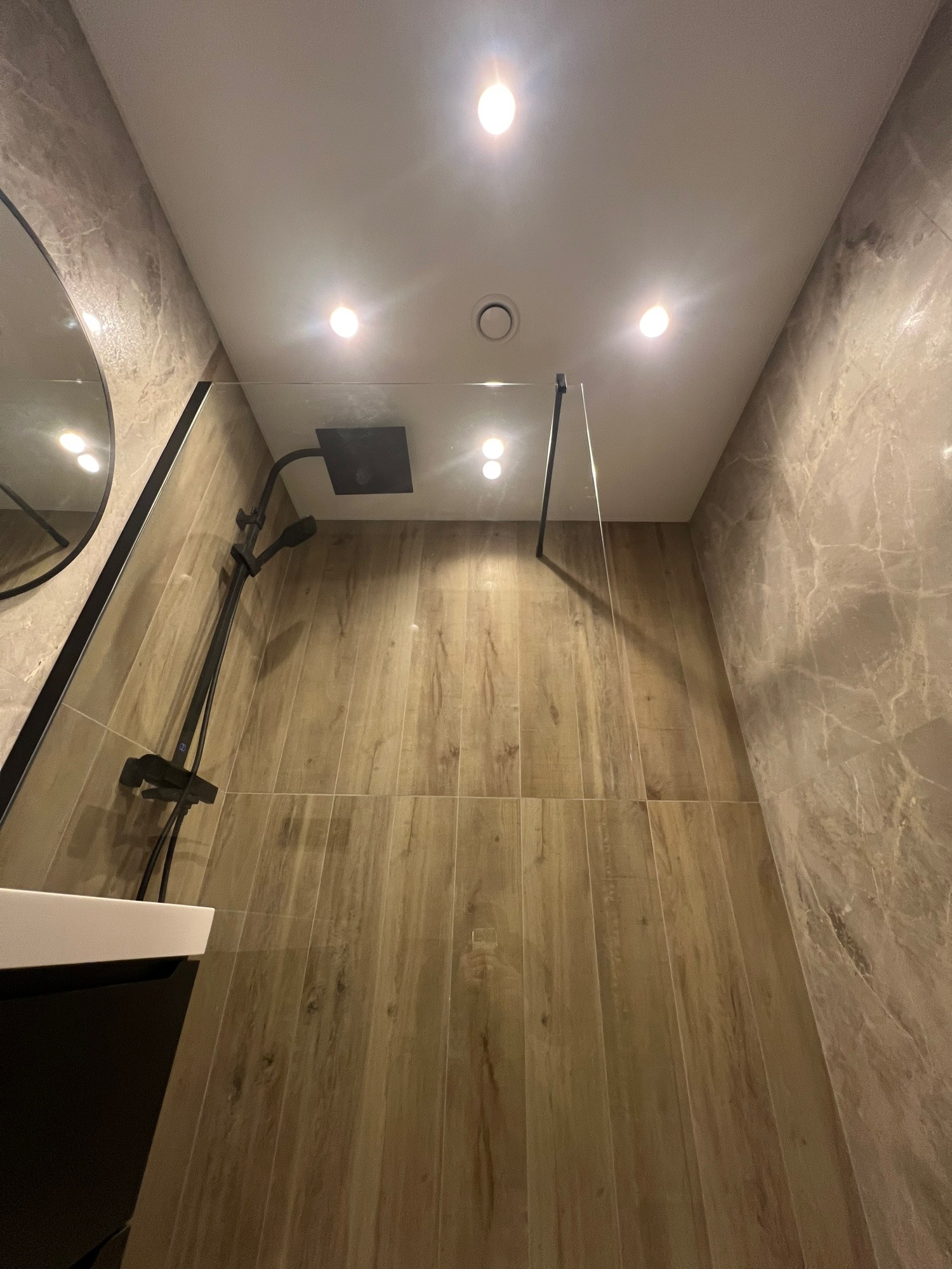 Nowoczesna kabina prysznicowa z czarną armaturą i szklaną ścianką, oświetlona punktowymi lampami. Płytki imitujące drewno na ścianach i podłodze. Minimalistyczny design.