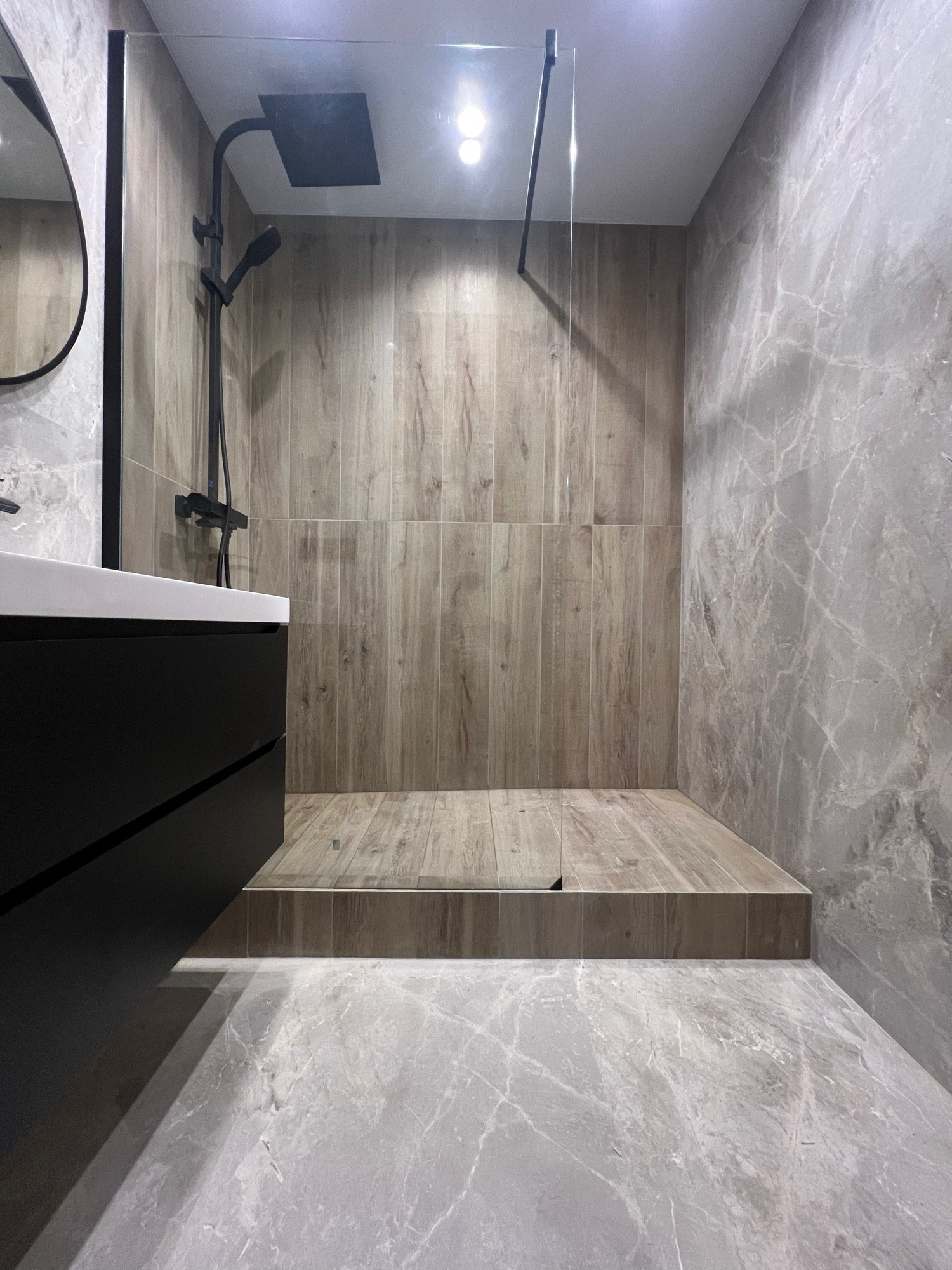 Nowoczesna łazienka z kabiną typu walk-in, wykończona płytkami imitującymi drewno i marmur, z czarną armaturą i szafką pod umywalką. Minimalistyczny design.