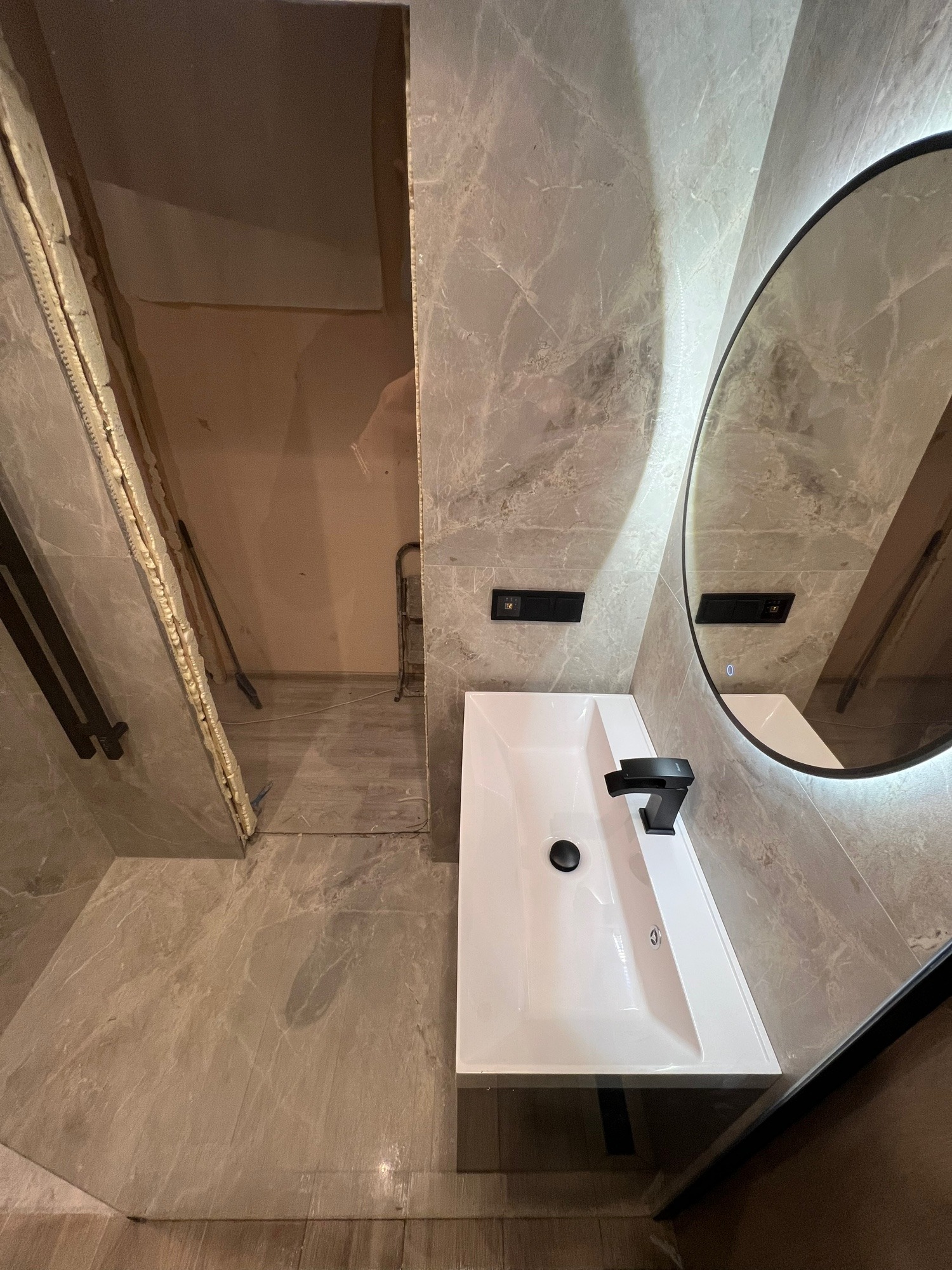 Łazienka w trakcie wykańczania: marmurowe płytki, biała umywalka z czarną armaturą, okrągłe lustro z podświetleniem i widoczne prace wykończeniowe w tle.