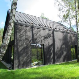 WOODIT Sp. z o.o. - Czarny, nowoczesny dom z drewnianą elewacją i metalowym dachem, otoczony zielenią i brzozami. Minimalistyczny design z dużymi oknami.