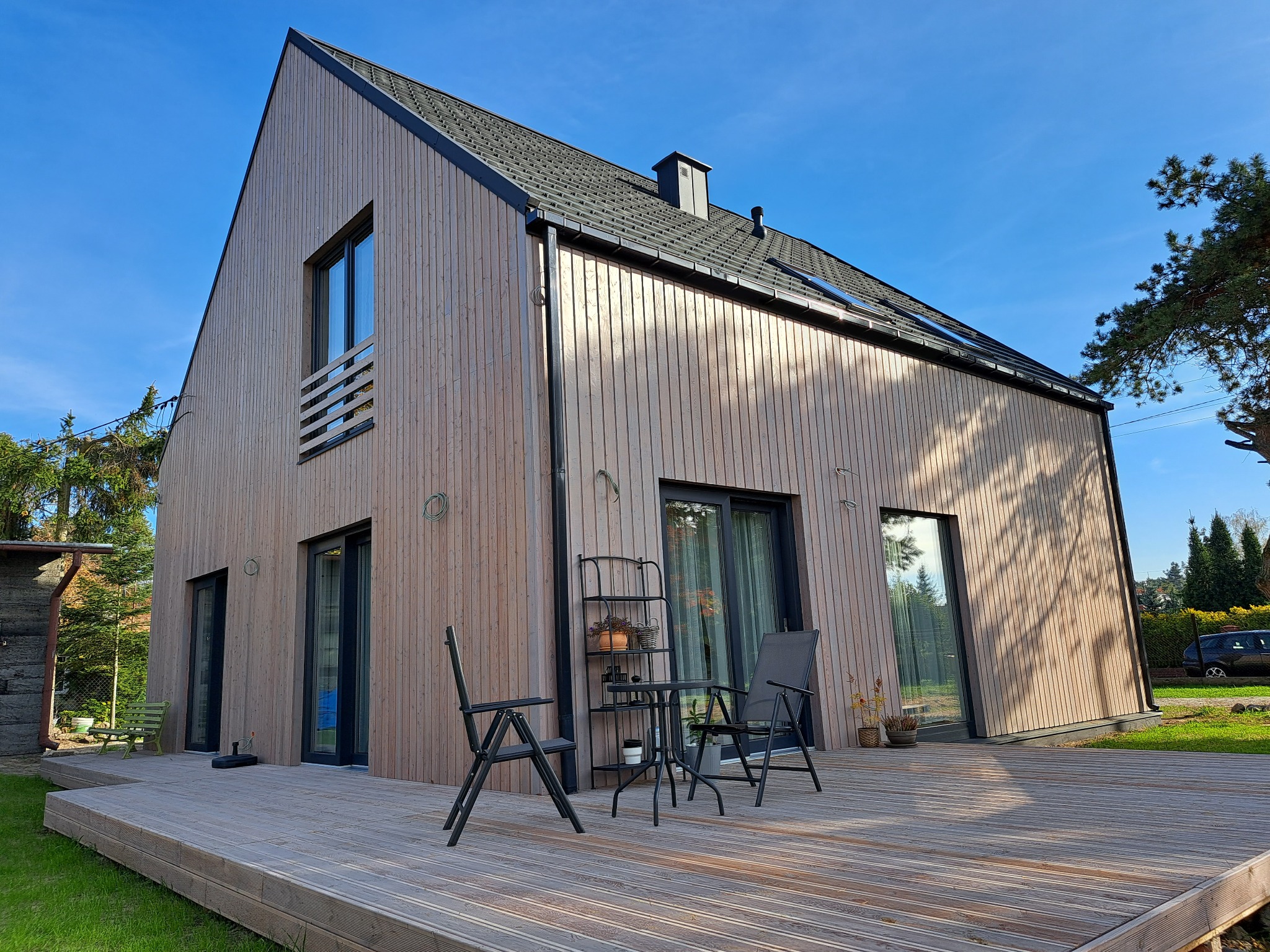 Nowoczesny dom z drewnianą elewacją i czarnym dachem, widok z tarasu z meblami ogrodowymi. Minimalistyczny design, jasne drewno, błękitne niebo w tle.