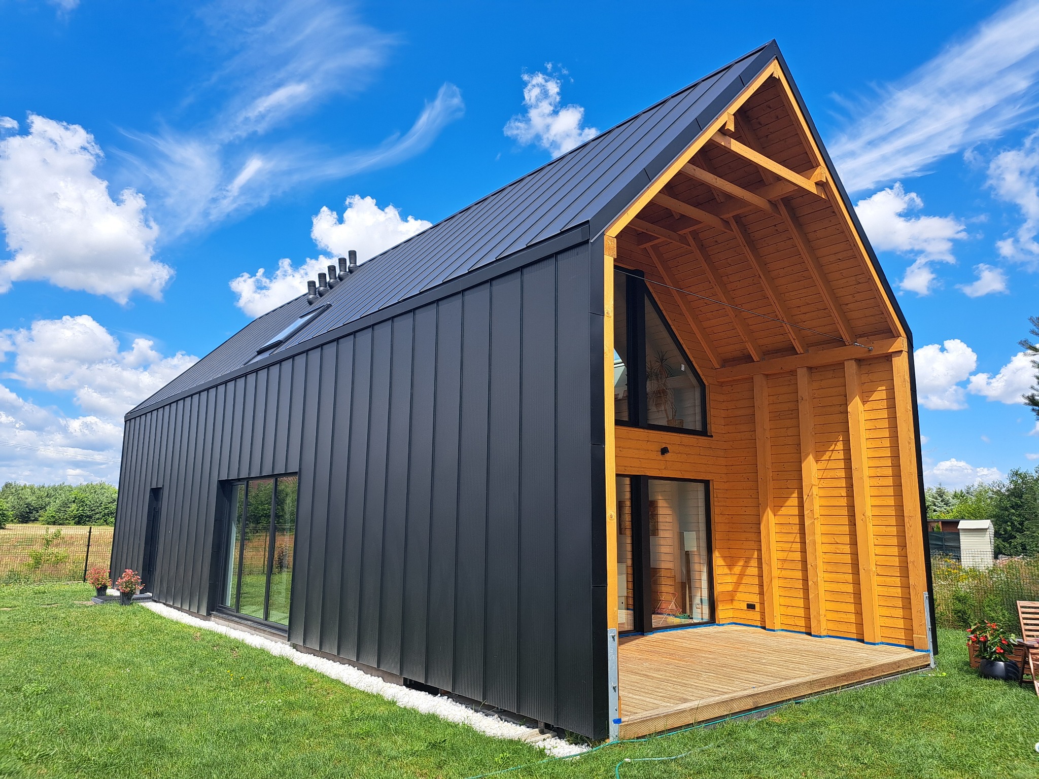 Nowoczesny dom z czarną elewacją i drewnianym szkieletem, widoczny taras i okna. Błękitne niebo z chmurami w tle. Ujęcie z poziomu trawnika.