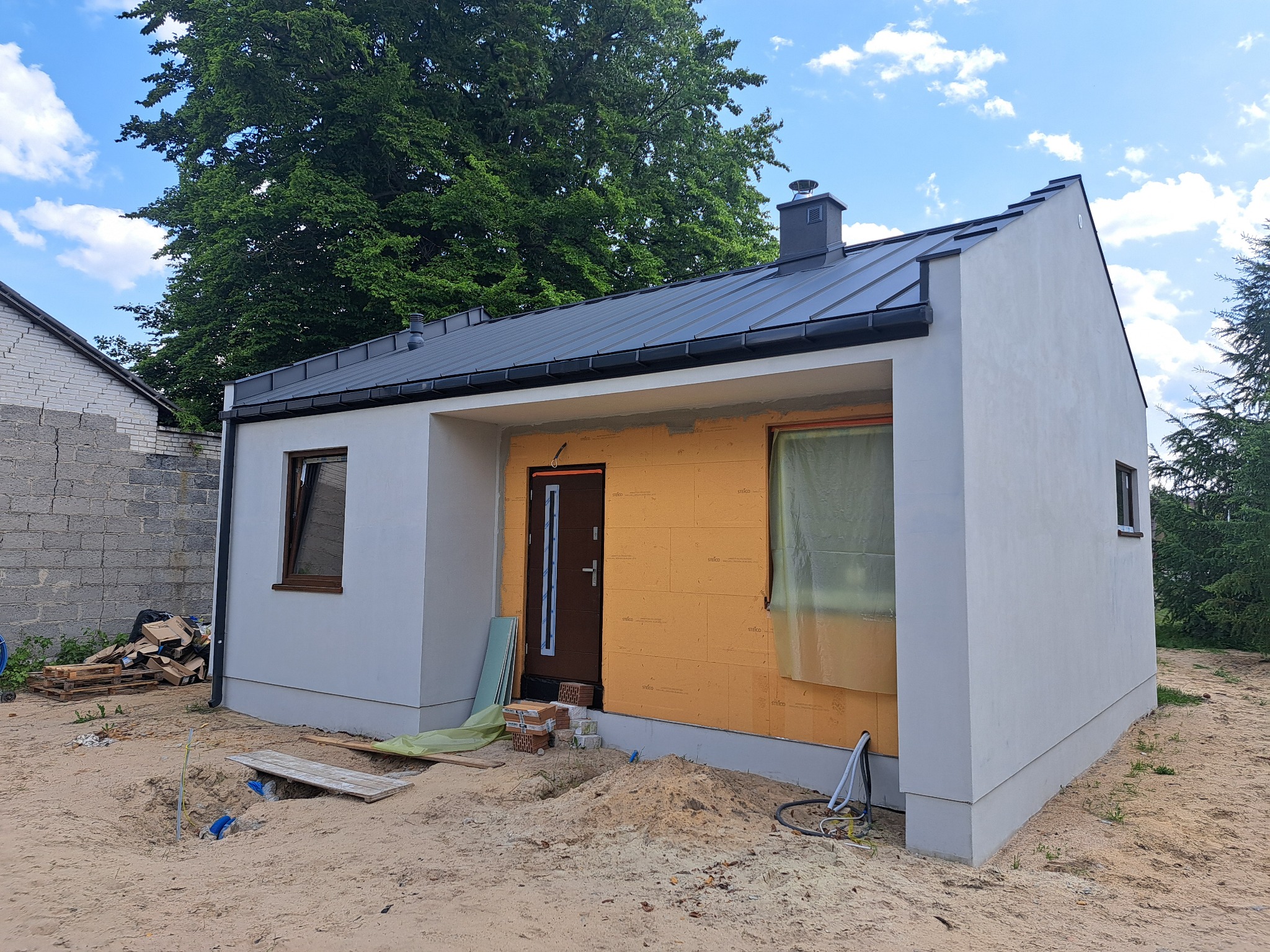 Niedokończony dom modułowy z szarą elewacją, czarnym dachem i oknami z brązowymi ramami. Widoczne ocieplenie ścian i przygotowania do dalszych prac wykończeniowych.