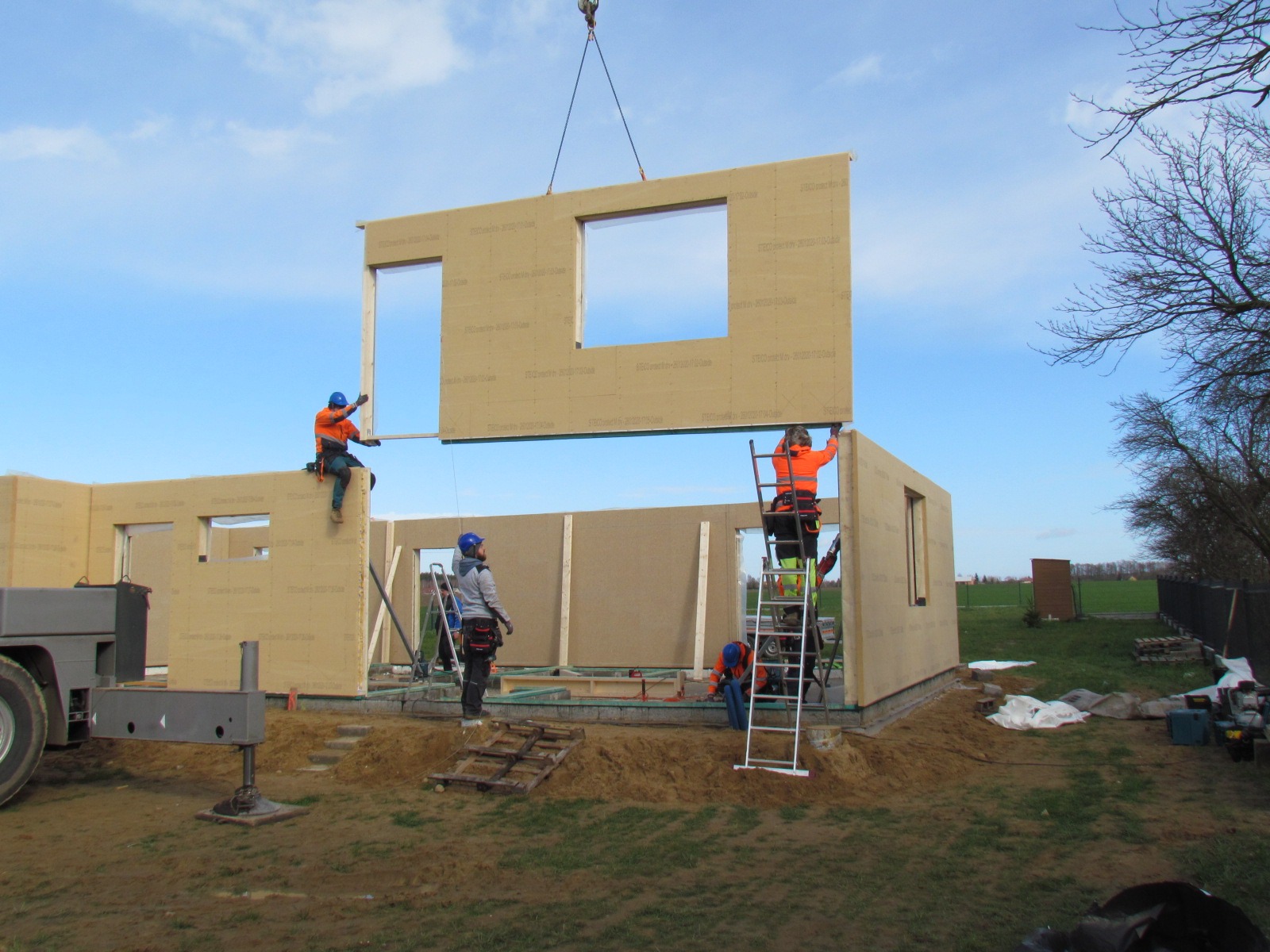 Montaż modułowej ściany domu z oknami za pomocą dźwigu. Pracownicy w kaskach i odzieży roboczej na budowie. Widoczne elementy konstrukcyjne i narzędzia.