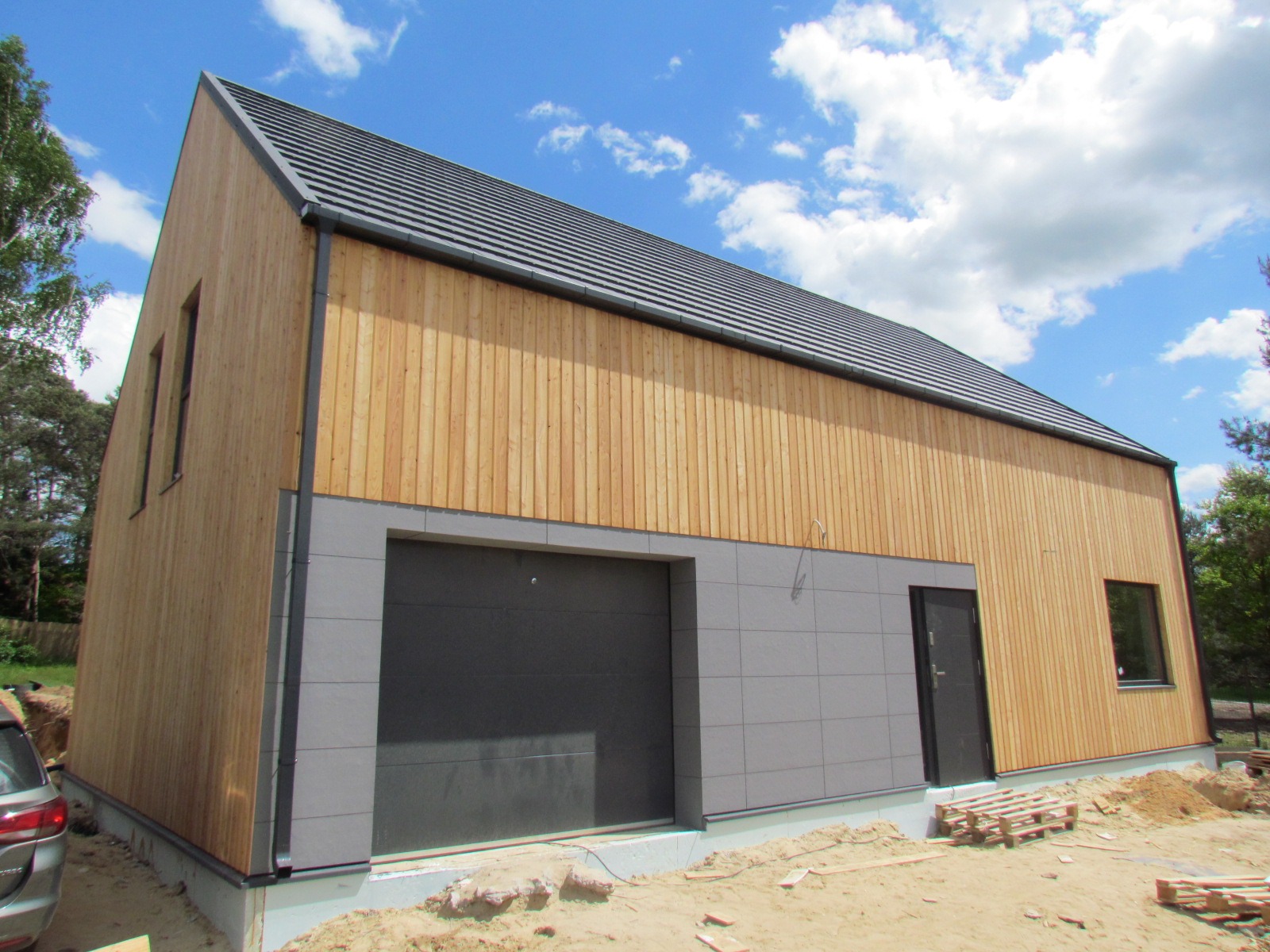 Nowoczesny dom z drewnianą elewacją i szarą częścią dolną, garaż z bramą segmentową, czarne okna i drzwi, dach pokryty szarą dachówką, błękitne niebo w tle.