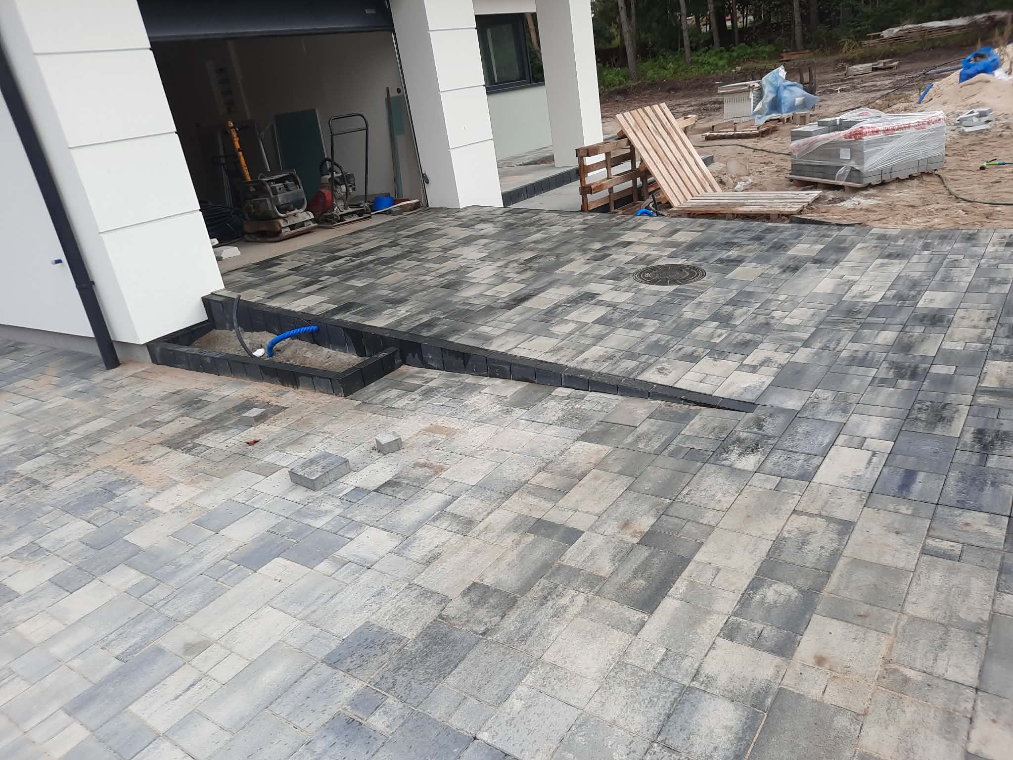 Ułożona, szara kostka brukowa przed domem z garażem. Widoczne palety z kostką i maszyny budowlane. Częściowo ułożona kostka przy schodach.