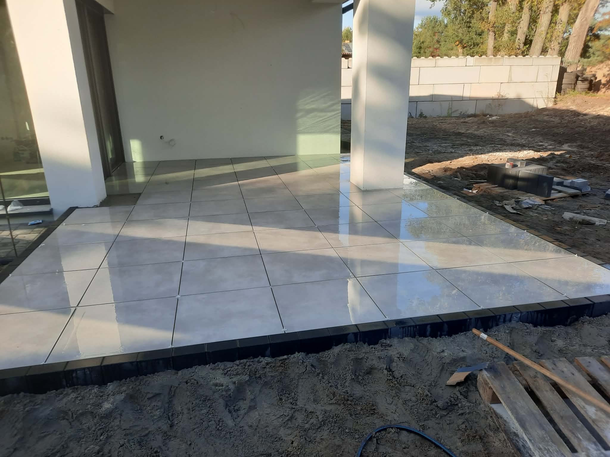 Jasne płytki tarasowe ułożone przy domu, obrzeża z czarnej kostki. Widoczne narzędzia i ziemia, co wskazuje na trwające prace wykończeniowe.