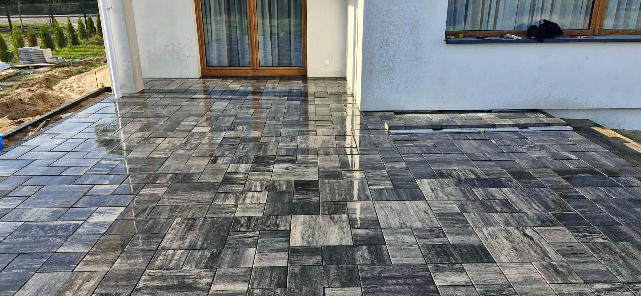 Nowoczesny taras z szarej kostki brukowej o nieregularnym wzorze, przylegający do białego budynku z drewnianymi oknami. Częściowo ułożona nawierzchnia z poziomicą.