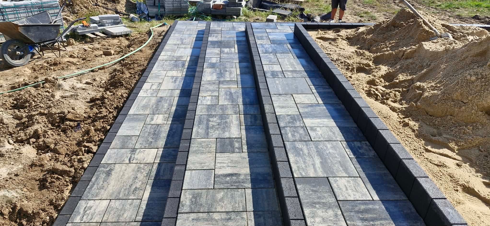 Ułożona ścieżka z kostki brukowej w odcieniach szarości, obrzeża z ciemnej kostki. W tle widoczna sterta piasku i narzędzia, co sugeruje trwające prace wykończeniowe.