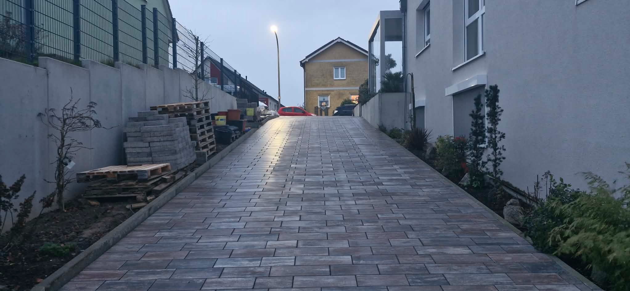 Ukończony podjazd z kostki brukowej w odcieniach szarości i brązu, prowadzący w górę w kierunku domów. Po lewej stronie palety z kostką i rosnące krzewy.