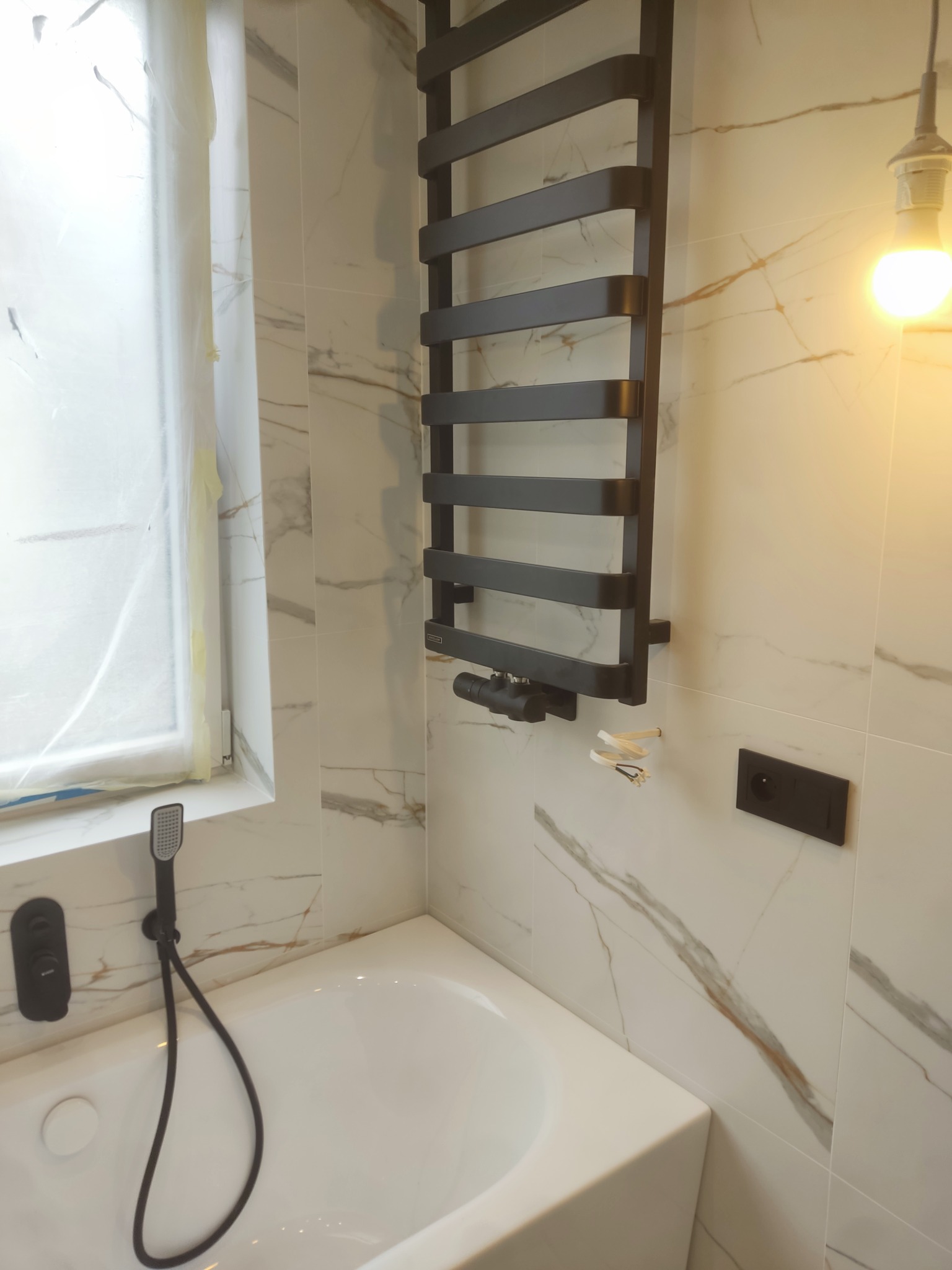 Nowoczesna łazienka z wanną, grzejnikiem drabinkowym i marmurowymi płytkami. Okno zabezpieczone folią malarską. Minimalistyczny design z czarnymi akcentami.