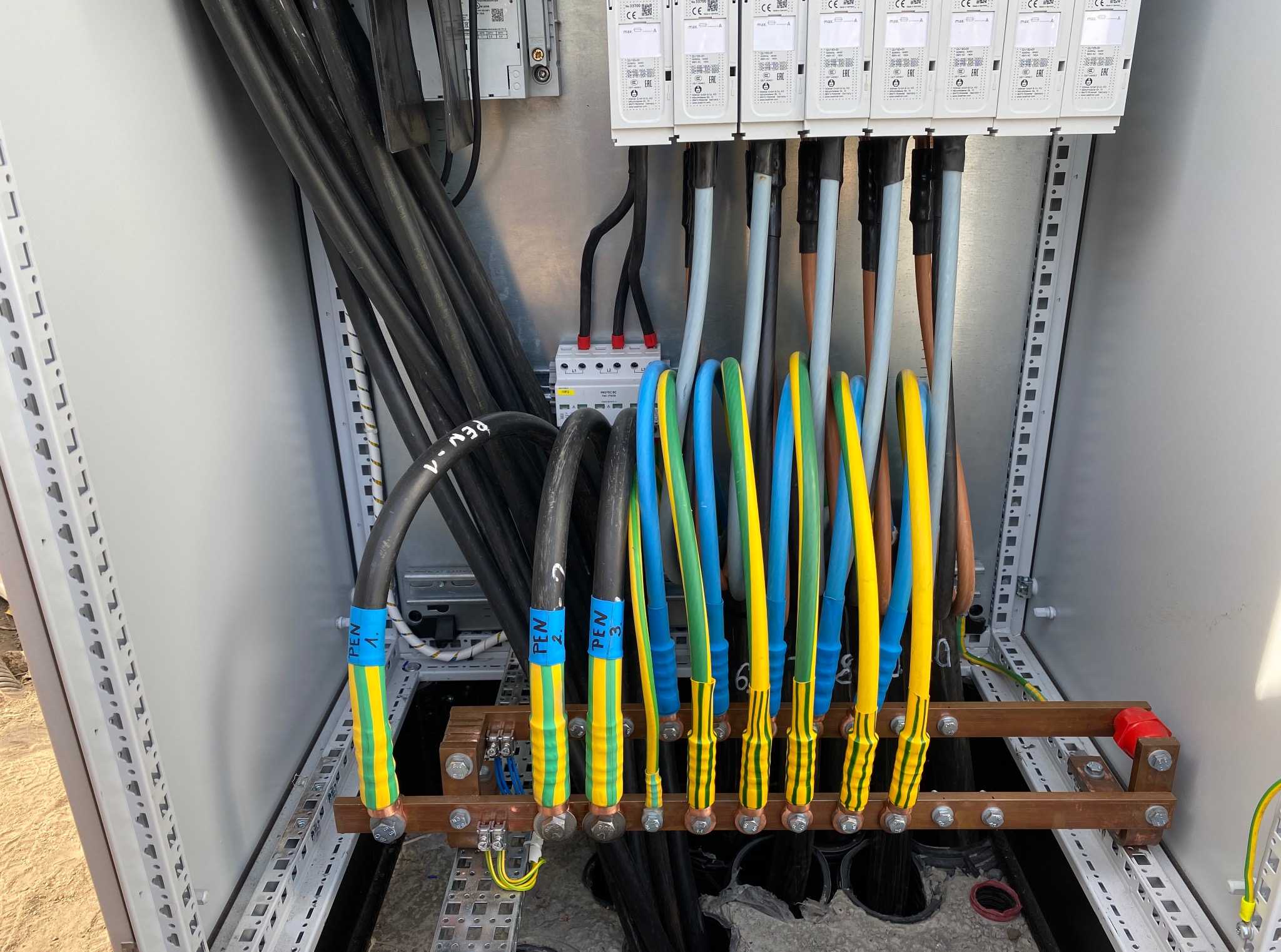 Wnętrze szafy elektrycznej z grubymi kablami oznaczonymi kolorowymi taśmami (żółto-zielono-niebieskimi) i podłączonymi do szyny uziemiającej. Widoczne zabezpieczenia nadprądowe.