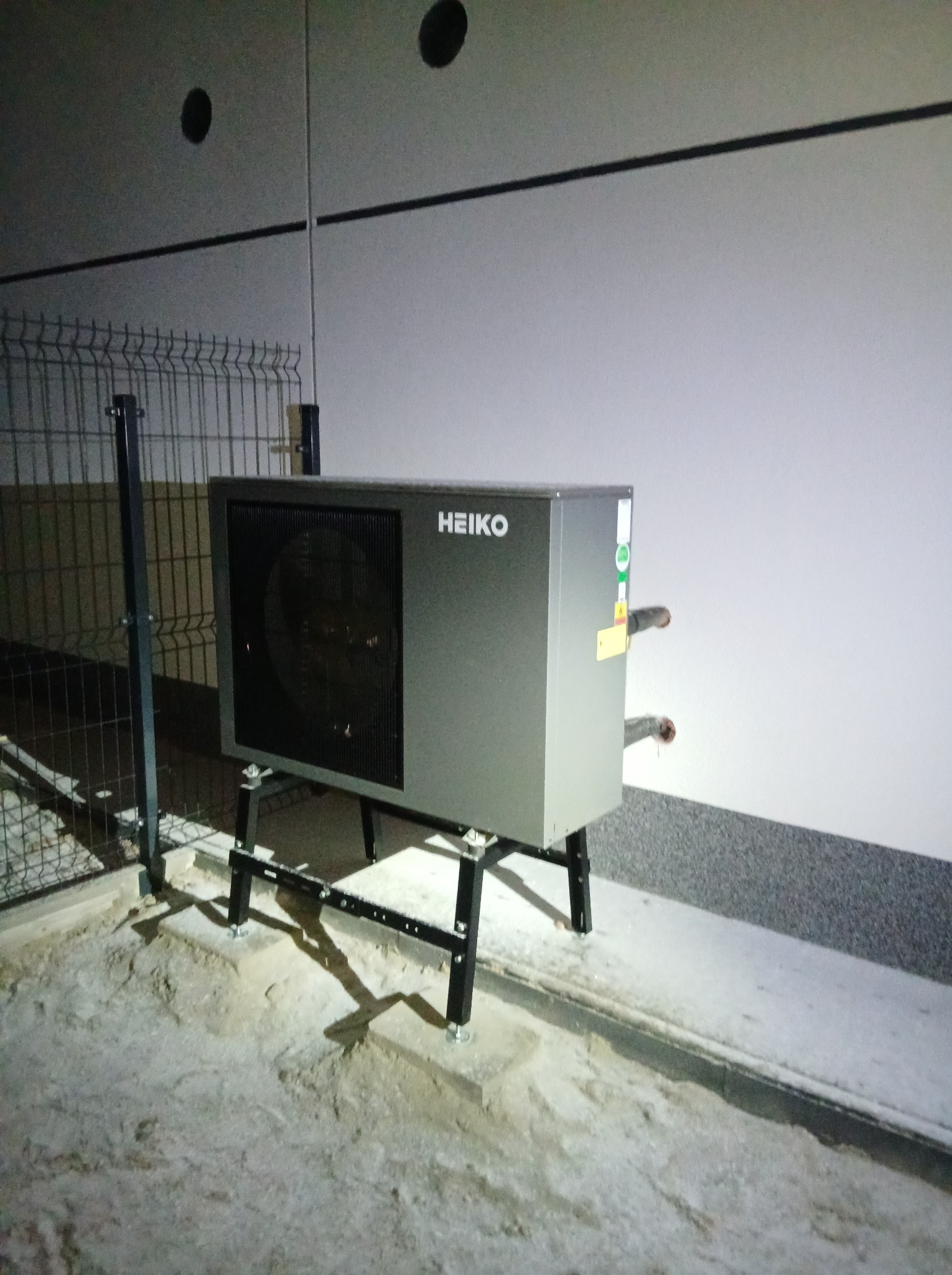 Szara pompa ciepła Heiko zamontowana na czarnym stelażu, widoczne rury i fragment ogrodzenia. Tło stanowi ściana z widocznymi otworami wentylacyjnymi.