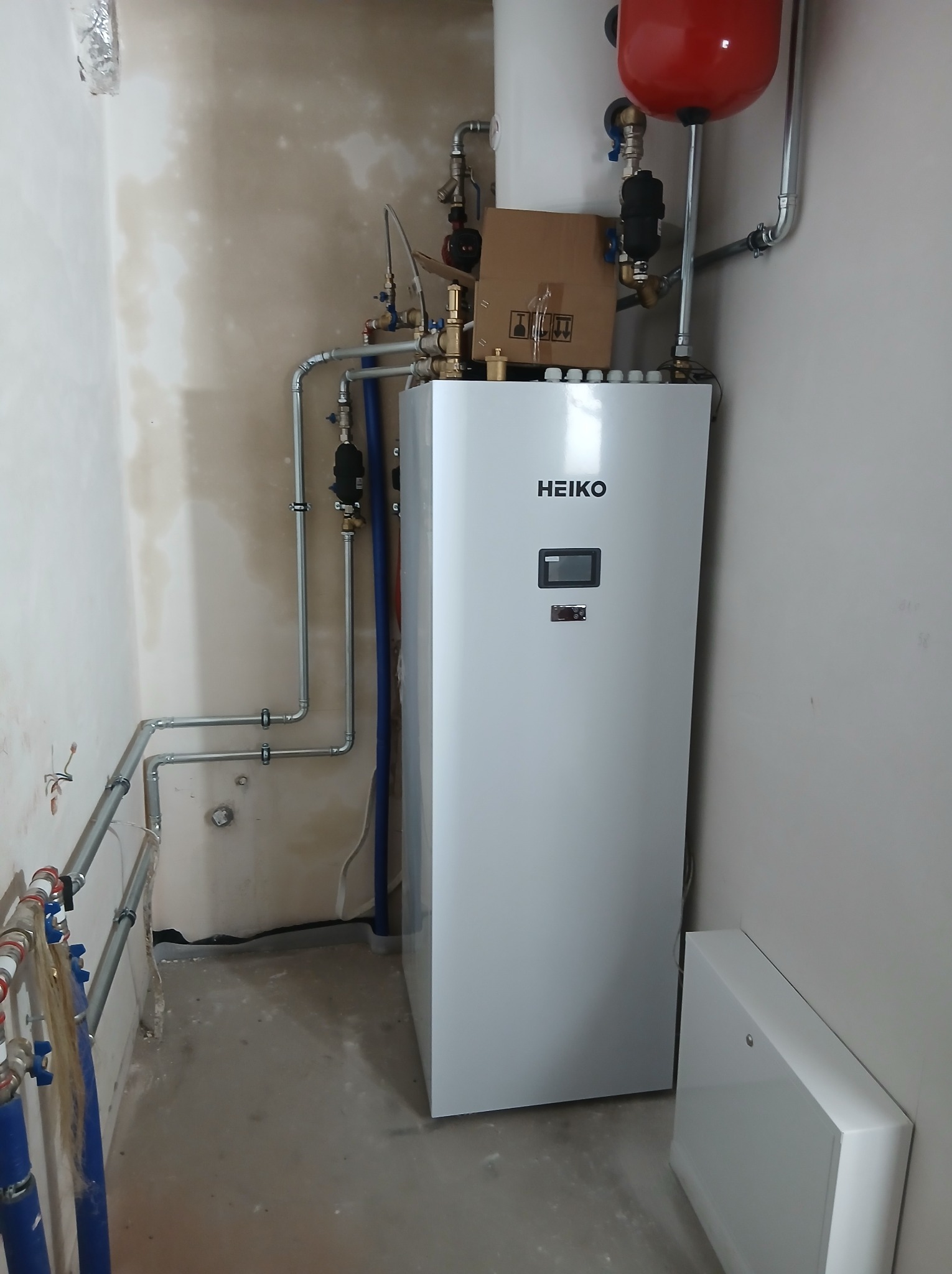 Biały piec HEIKO z podłączonymi rurami w surowym pomieszczeniu. Widoczny zbiornik wyrównawczy i skrzynka. Instalacja w trakcie prac wykończeniowych.