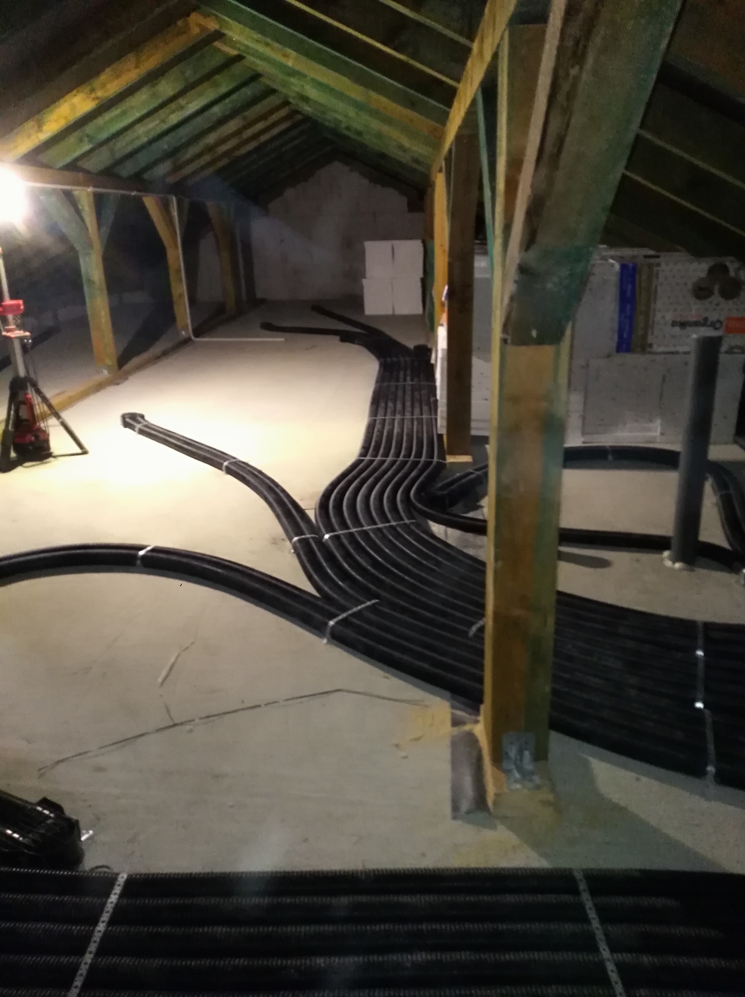 Czarny system rur rekuperacji ułożony na poddaszu z drewnianą konstrukcją. Widoczny fragment oświetlenia roboczego.