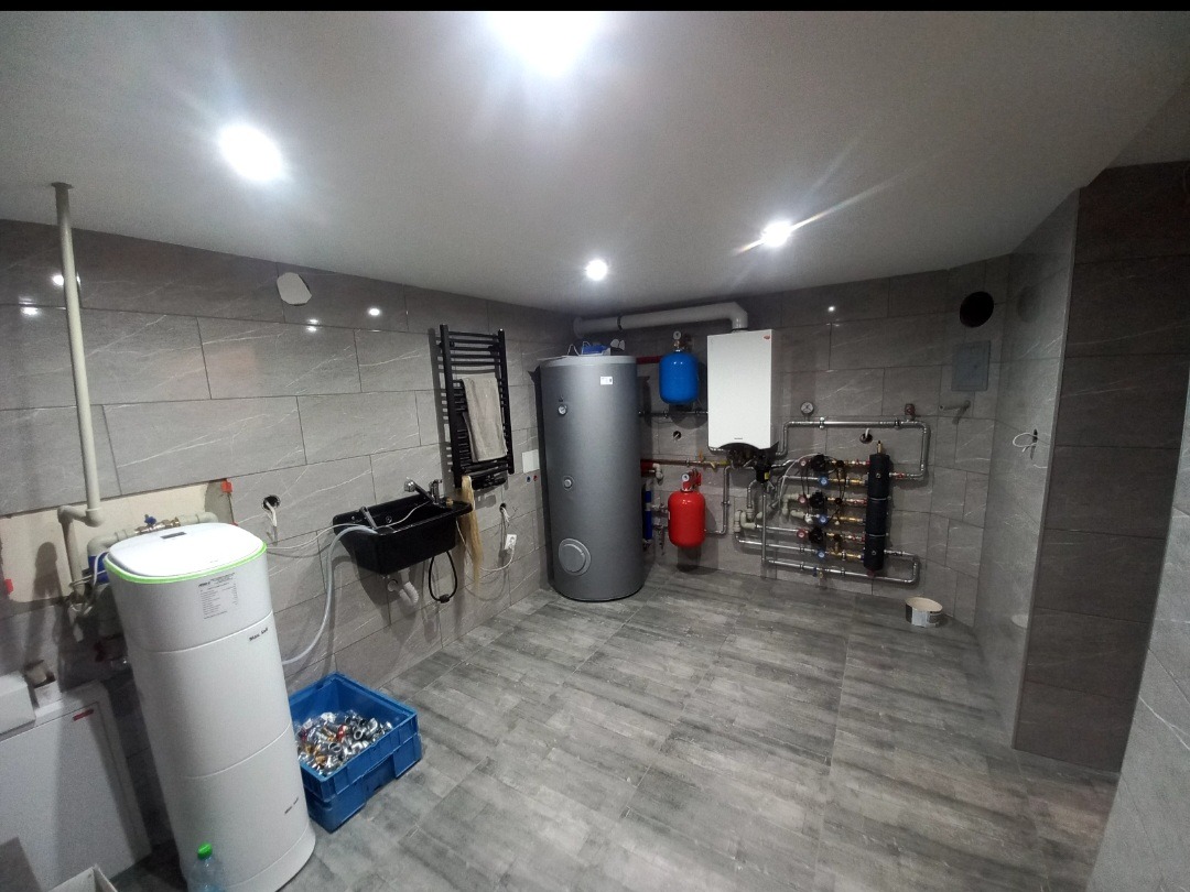 Nowoczesna kotłownia z szarymi płytkami, zbiornikiem, piecem gazowym, grzejnikiem drabinkowym i instalacją hydrauliczną. Widoczny pojemnik z elementami instalacyjnymi.