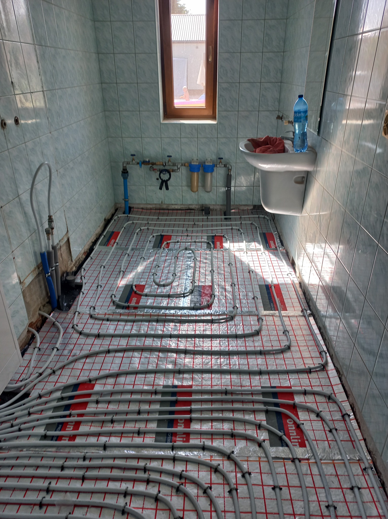 Instalacja ogrzewania podłogowego w łazience z jasnymi płytkami. Widoczne rury na folii izolacyjnej z czerwonym nadrukiem, armatura i umywalka.
