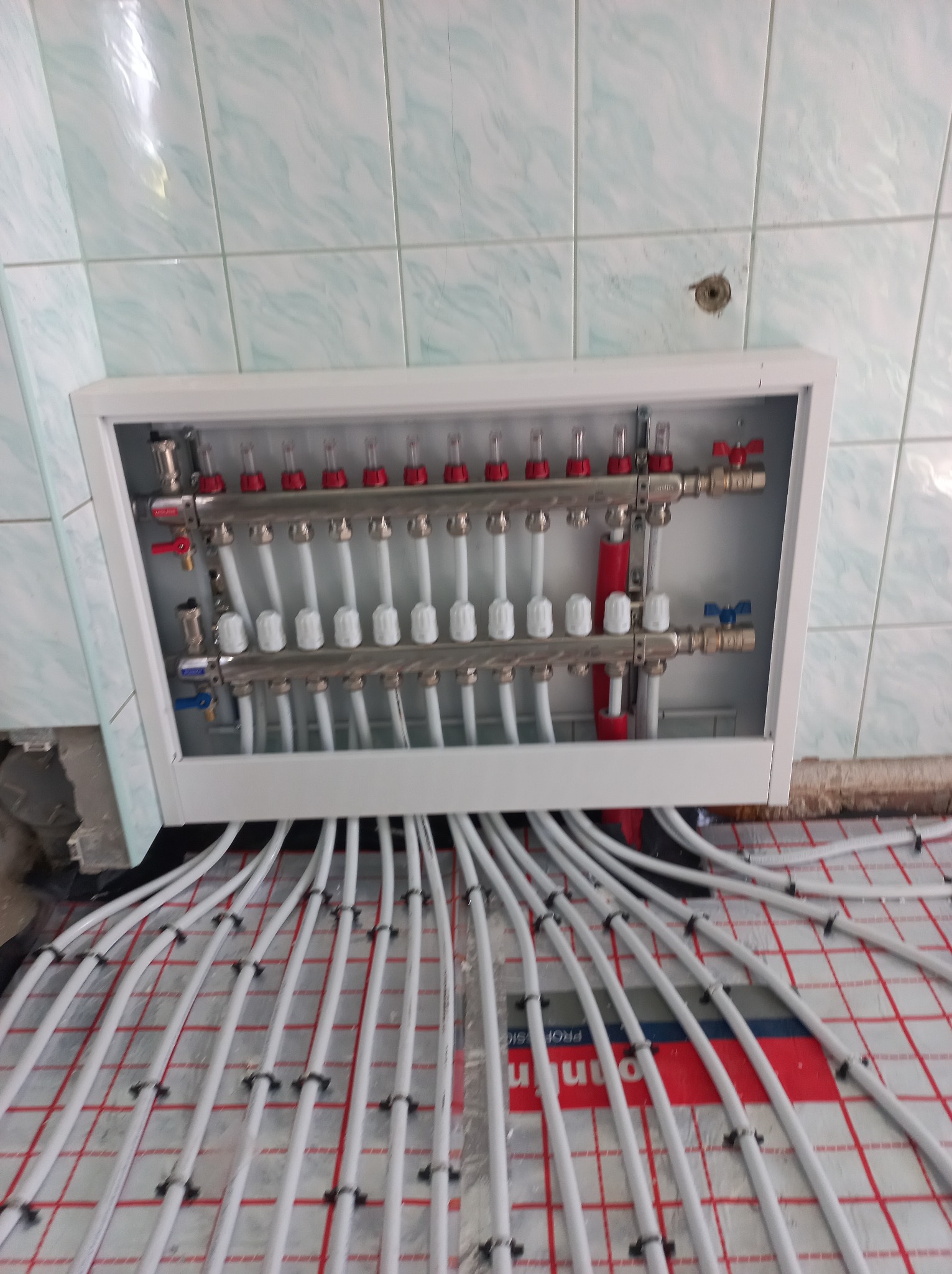 Rozdzielacz ogrzewania podłogowego w białej obudowie, z widocznymi rurami na podkładzie z siatką. Ujęcie z góry, widoczne płytki na ścianie.