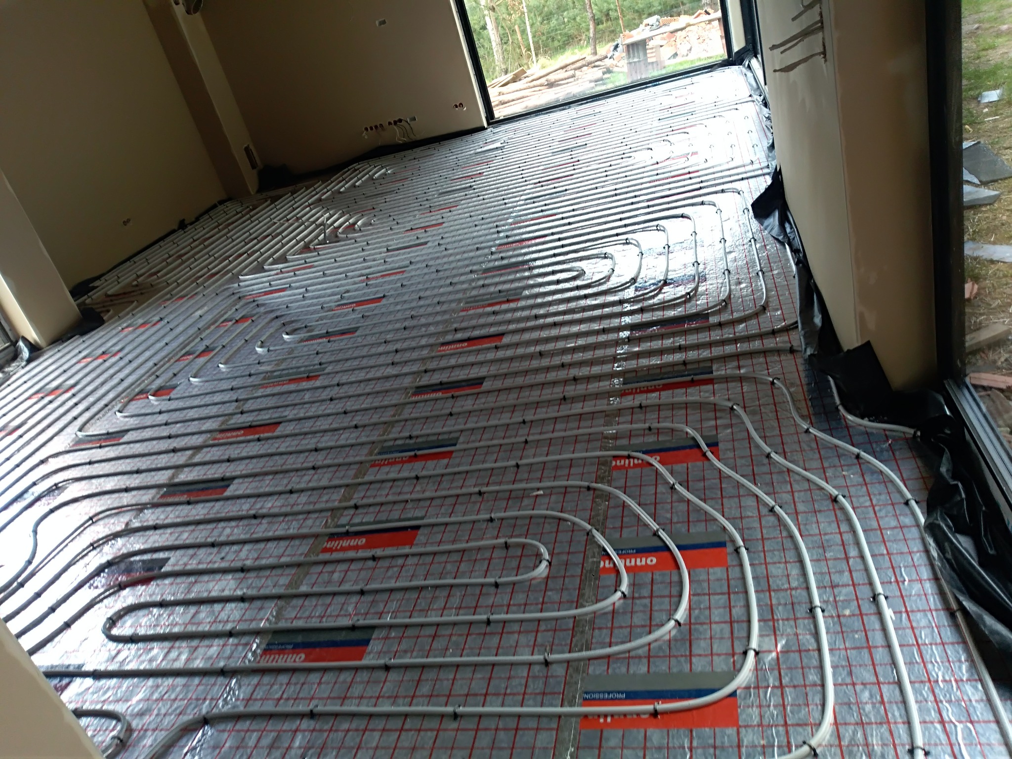 Ułożone rury ogrzewania podłogowego na folii izolacyjnej z widocznymi mocowaniami, przygotowane do zalania posadzką w nowym budynku z dużym oknem.