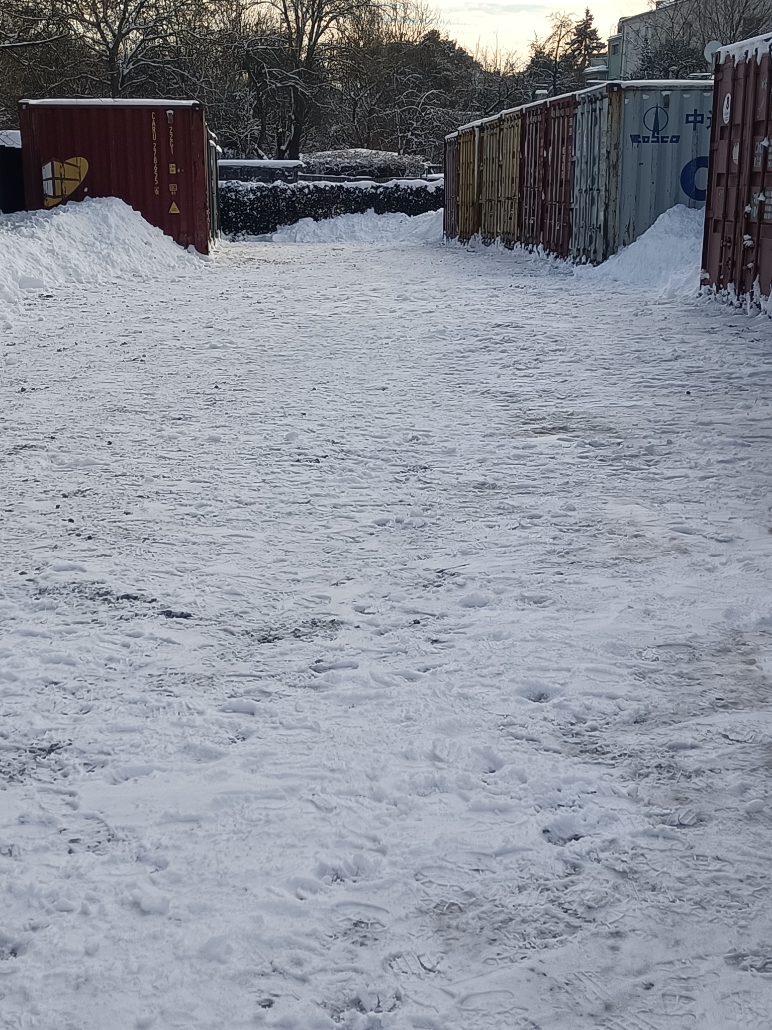 Zasypany śniegiem plac z rzędem kontenerów w tle. Widoczne ślady stóp na śniegu. Zimowy krajobraz z kontenerami.
